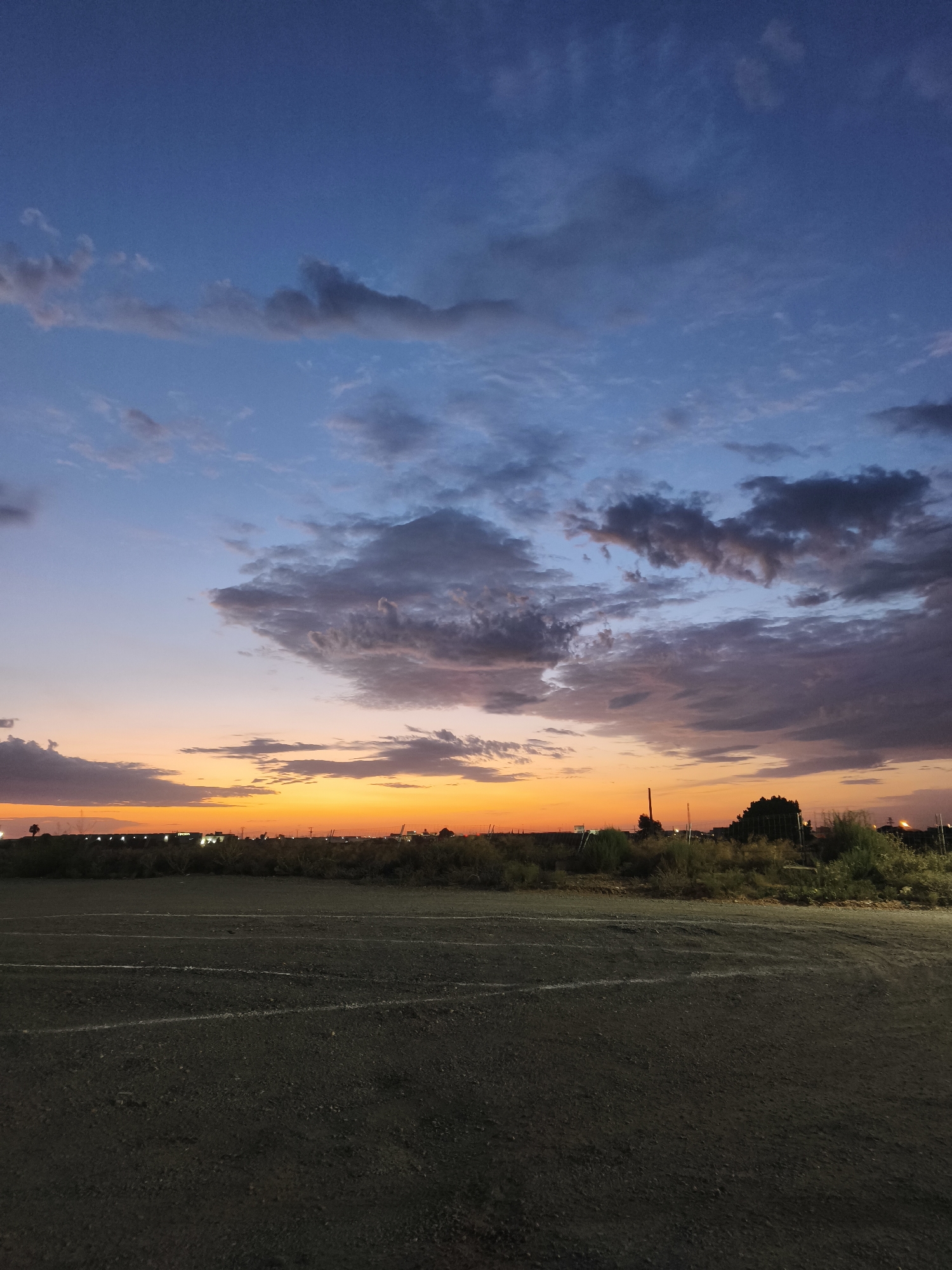 The image captures a dusky sky over a semi-arid landscape, likely in or near Picassent, Spain. The sky transitions from a deep blue at the top to vibrant orange and yellow hues near the horizon, indicative of a sunset or sunrise. Scattered clouds, tinged with purple and grey, drift across the sky. In the middle ground, a flat, open area with some vegetation and a few distant structures, possibly industrial or agricultural buildings, are visible. The foreground consists of a dark, rough-textured ground surface, possibly a dirt or gravel lot, marked with faint white lines. The overall impression is one of a rural or industrial outskirts setting during twilight.