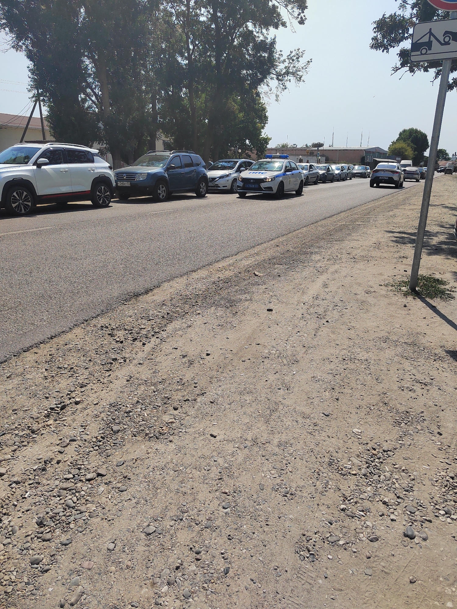 A traffic jam is occurring on a paved road in Vityazevo, Russia. Several vehicles, including a white SUV, a blue SUV, and a police car with flashing lights, are stopped in traffic. The road is lined with trees and buildings. The weather is sunny and clear. The ground beside the road is unpaved and covered with gravel and sand. A traffic sign indicating a tow truck is visible on the right side of the image.