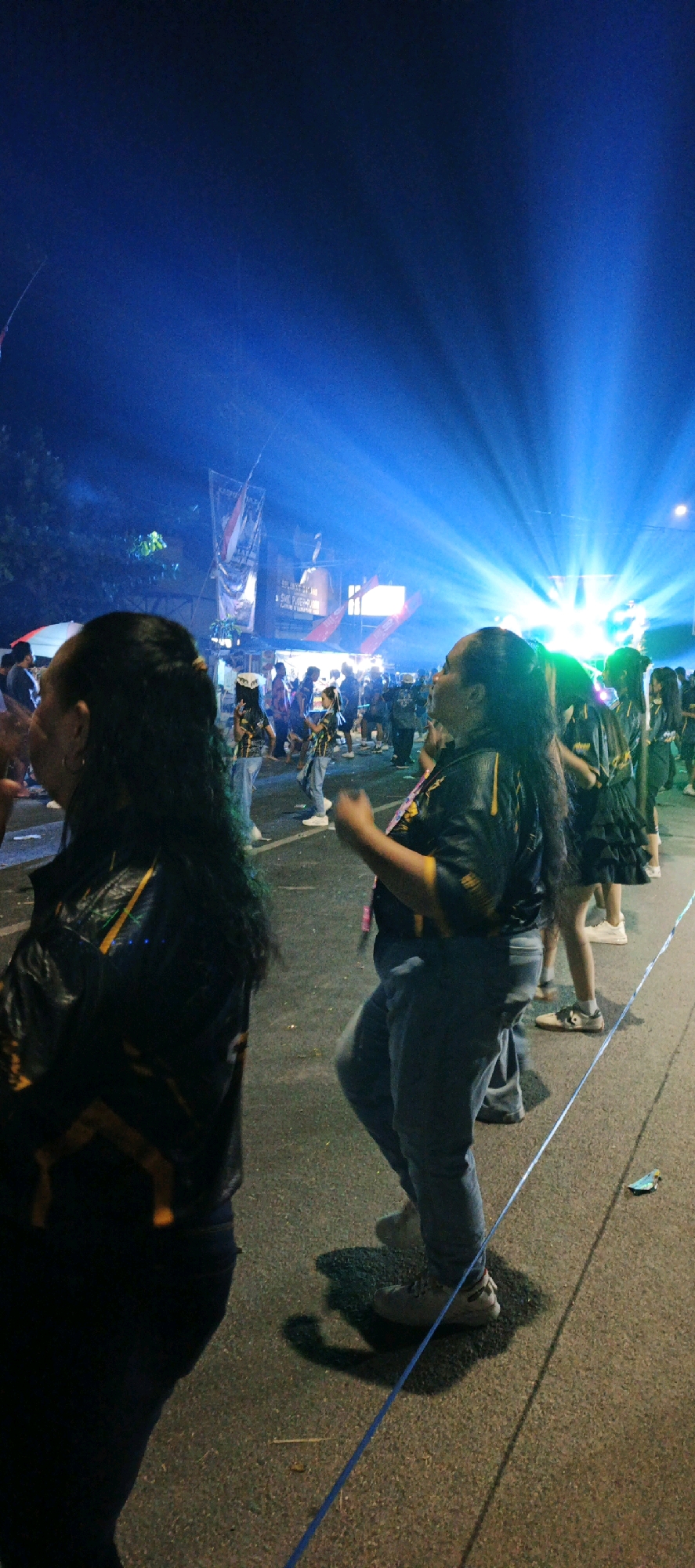 The image captures an outdoor nighttime scene in the city of Kasiyan, Indonesia, likely during a festival or event. Bright blue spotlights illuminate the sky from the background, creating dramatic rays of light. In the foreground, several people are visible. To the left, a person with dark hair is seen from behind, wearing a black jacket with yellow accents. To the right of center, another person, also with dark hair, is in motion, appearing to dance or celebrate. This individual is wearing a black and yellow shirt and grey pants. In the background, a crowd of people can be seen, along with banners and structures that suggest a street market or performance area. The ground is asphalt, with some debris visible. A thin blue ribbon or string runs diagonally across the lower portion of the image. The atmosphere appears lively and festive.