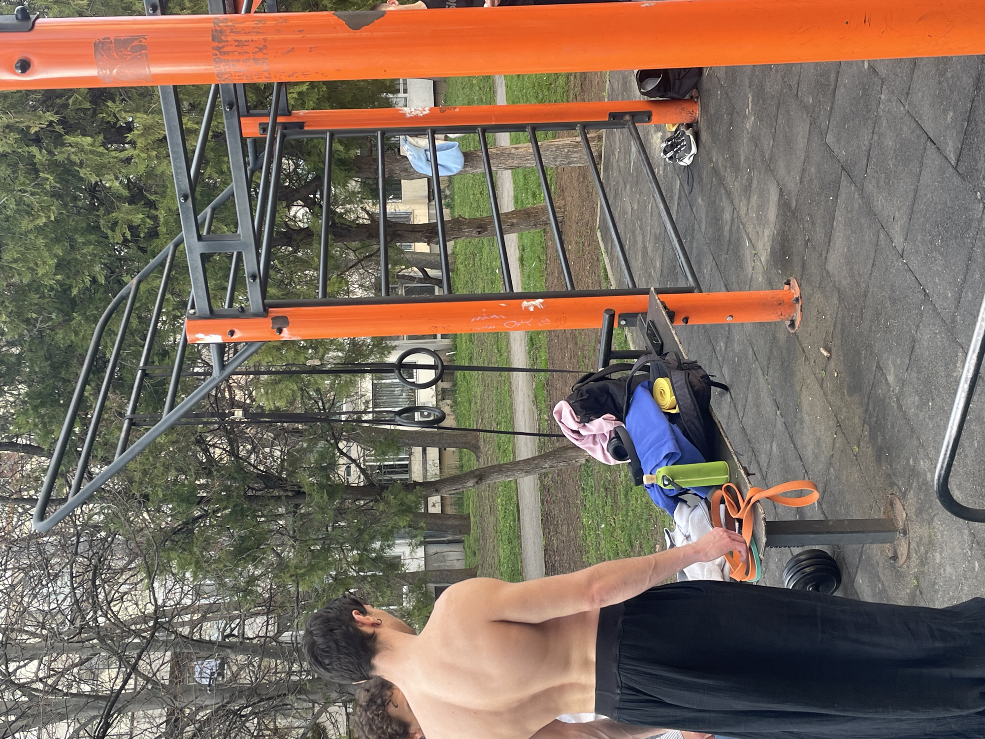 The image shows an outdoor exercise area. An orange and grey jungle gym-style structure dominates the midground. To the right of the jungle gym, there is exercise equipment, including a backpack with visible blue and yellow items, a green water bottle, and orange resistance bands. A shirtless man with dark hair and black pants is in the foreground. The background includes trees and buildings, suggesting an urban park. The location is likely in Sofia, Bulgaria.