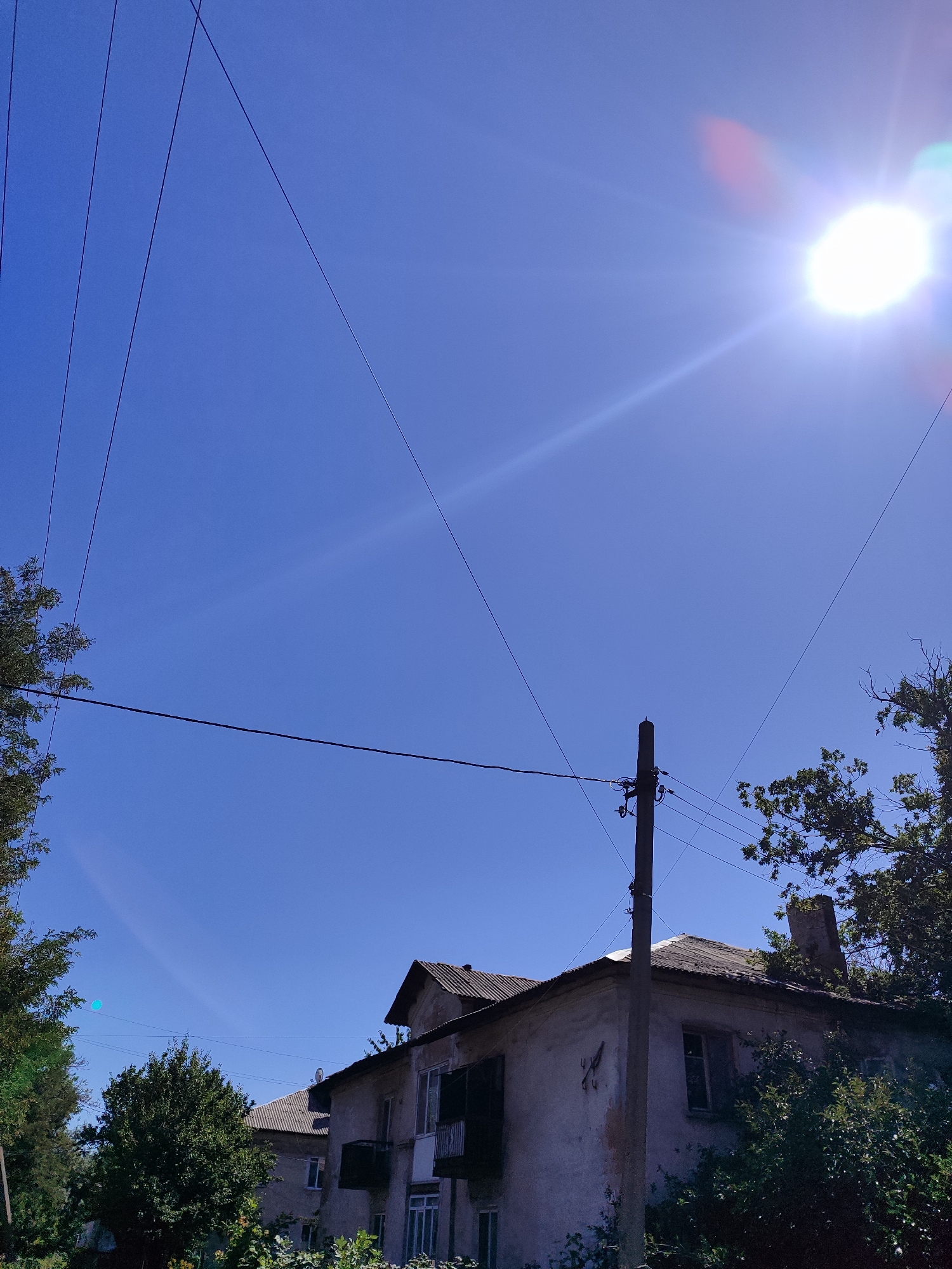 A sunny, clear blue sky dominates the upper portion of the image, with the sun appearing as a bright, overexposed orb in the top right quadrant, casting lens flares and a distinct sunbeam across the sky. Multiple thin, dark power lines stretch across the sky at various angles, some originating from a weathered concrete utility pole on the right side of the frame.
In the lower half of the image, an old, two-story building with a gray, weathered roof is partially visible. The building appears to be in disrepair, with peeling paint on its exterior walls and dark, shuttered windows. A small balcony with a railing is visible on the second floor. Adjacent to the building, dense green trees and foliage obscure parts of the structure and fill the foreground and right side of the image. Another building, similar in style but less visible, is partially seen behind the main structure. The scene appears to be an outdoor urban or suburban setting.