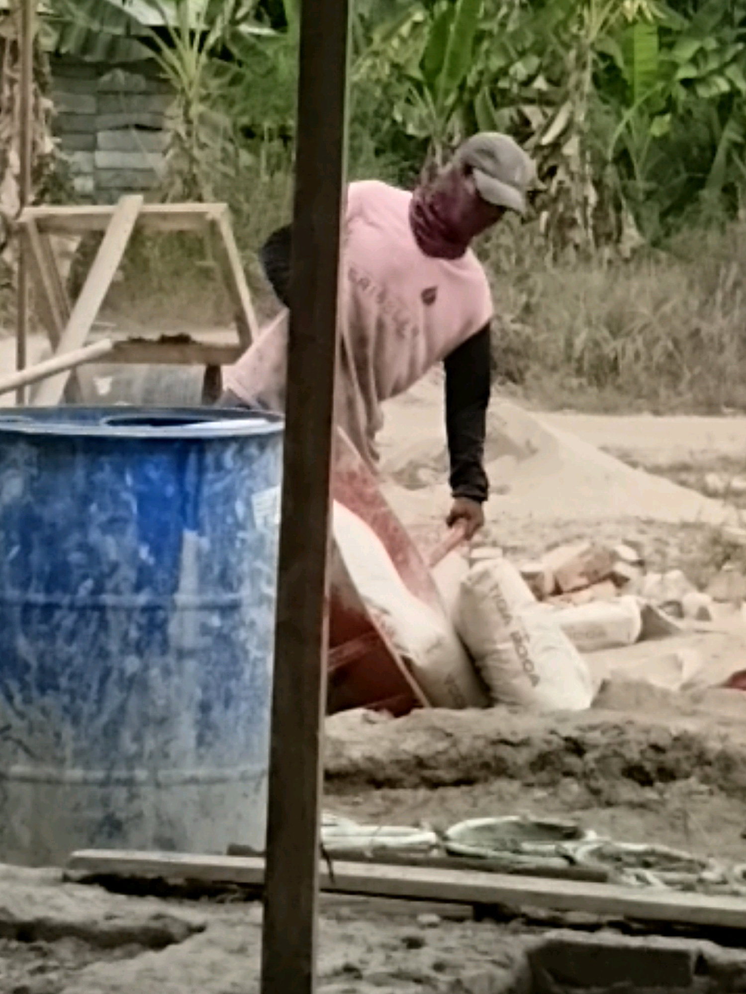 The image depicts a construction site in Margo Rukun, Indonesia. A man wearing a pink shirt and a grey cap is visible. He appears to be working with materials, possibly cement or sand, as several bags are laid out nearby. A large blue barrel is also present on the site. The environment suggests an outdoor setting with vegetation in the background. The overall scene indicates ongoing construction work.