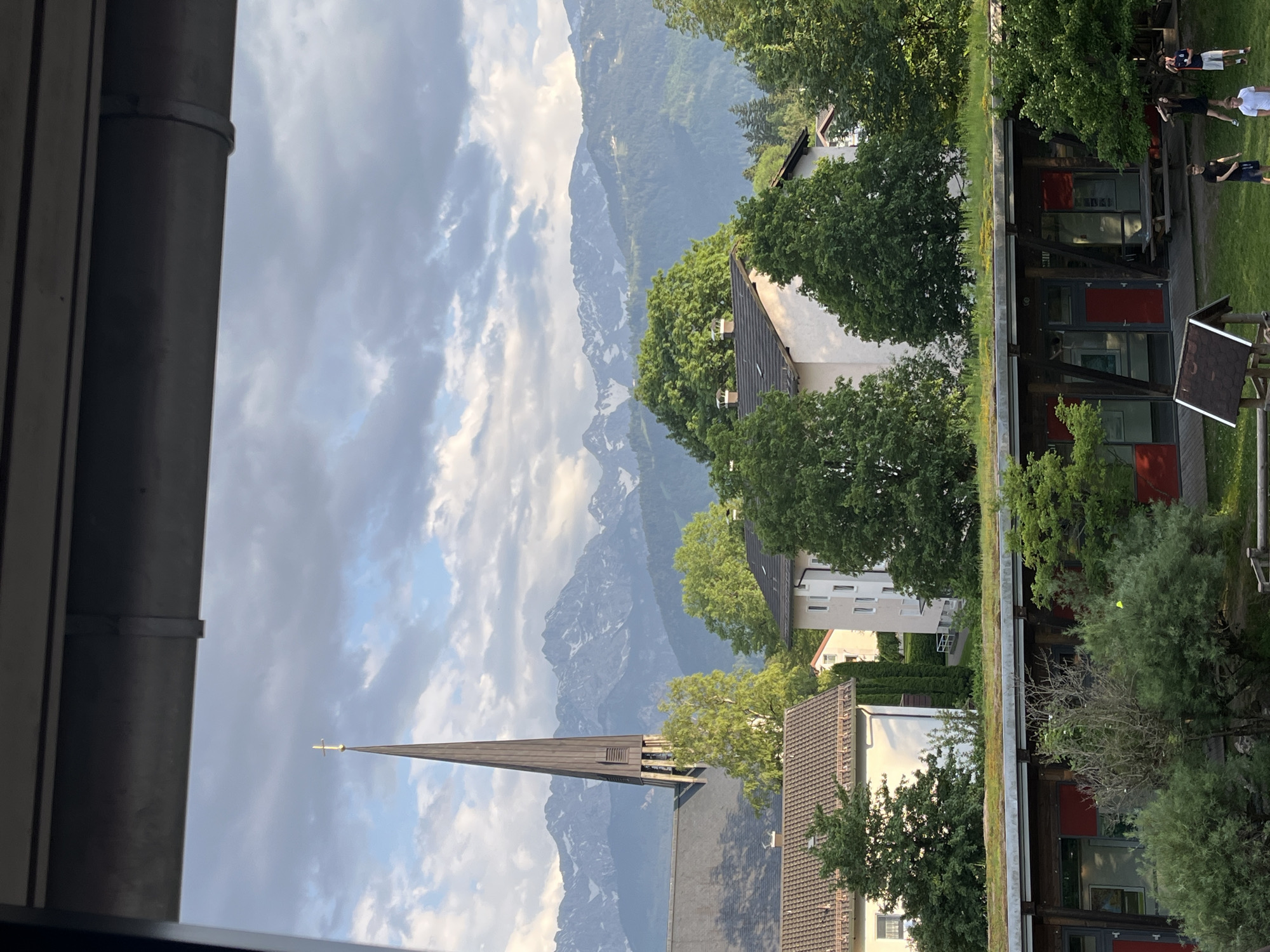 The image shows a partial view of Garmisch-Partenkirchen, Germany. In the foreground is part of a building, including a drainpipe. In the midground, there is a church steeple pointing towards a mountainous landscape with snow-capped peaks visible through the clouds. There are residential buildings partially hidden by trees. In the background there is a green lawn with a wooden building and a group of people. Some are sitting and some are standing in a group.