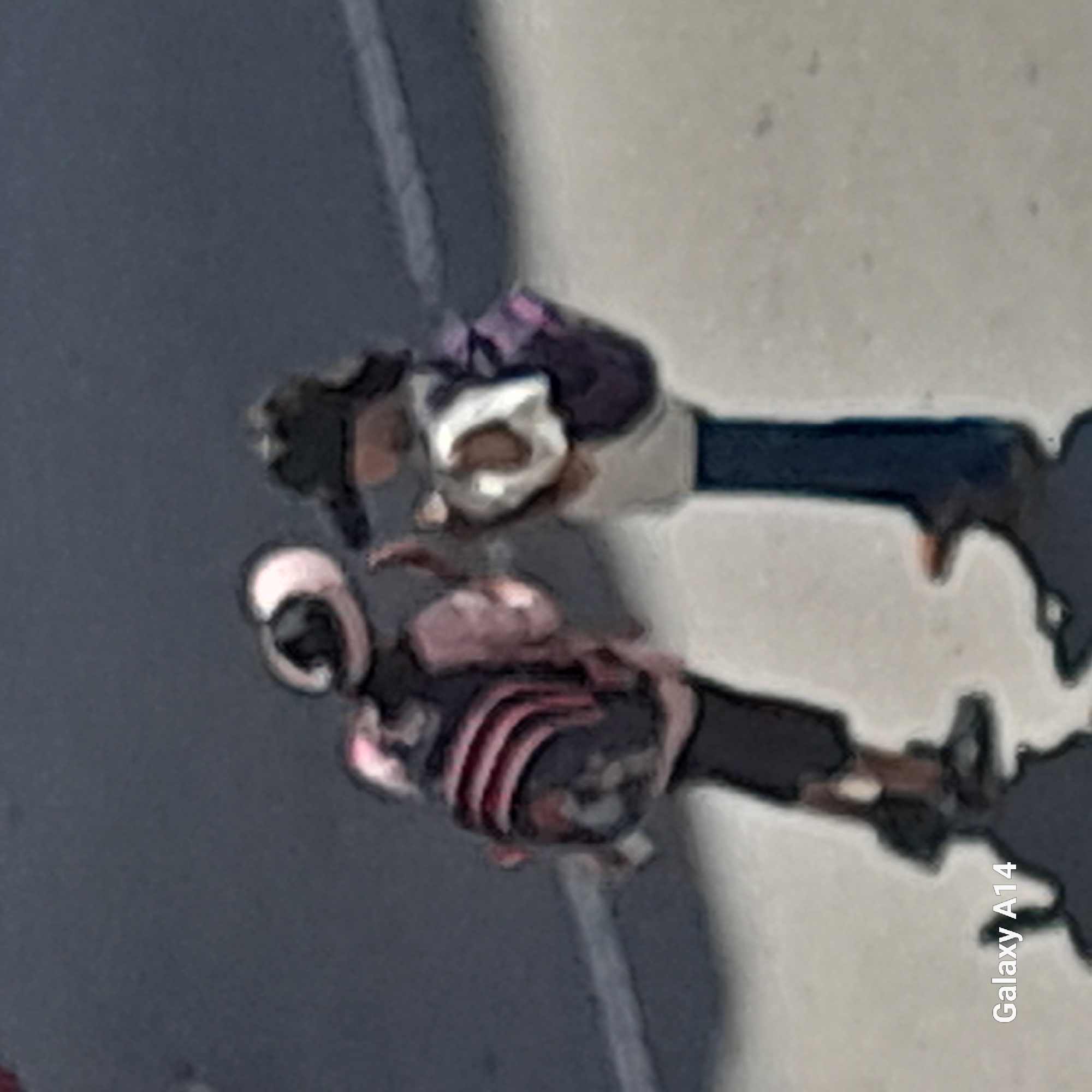 This is an out-of-focus, high-angle photograph taken in Khan Yunis, Palestinian Territories, showing two young individuals with backpacks facing each other. The person on the left wears a light-colored hat and a backpack with pink and black stripes. The person on the right wears a dark hat and a backpack that appears to be dark purple with a white element on the front. They are standing on a grey paved surface that transitions to a lighter, textured surface, possibly a sidewalk. The background is a dark, solid surface. The word "Galaxy A14" is visible in the bottom left corner of the image, indicating the device used for capture.