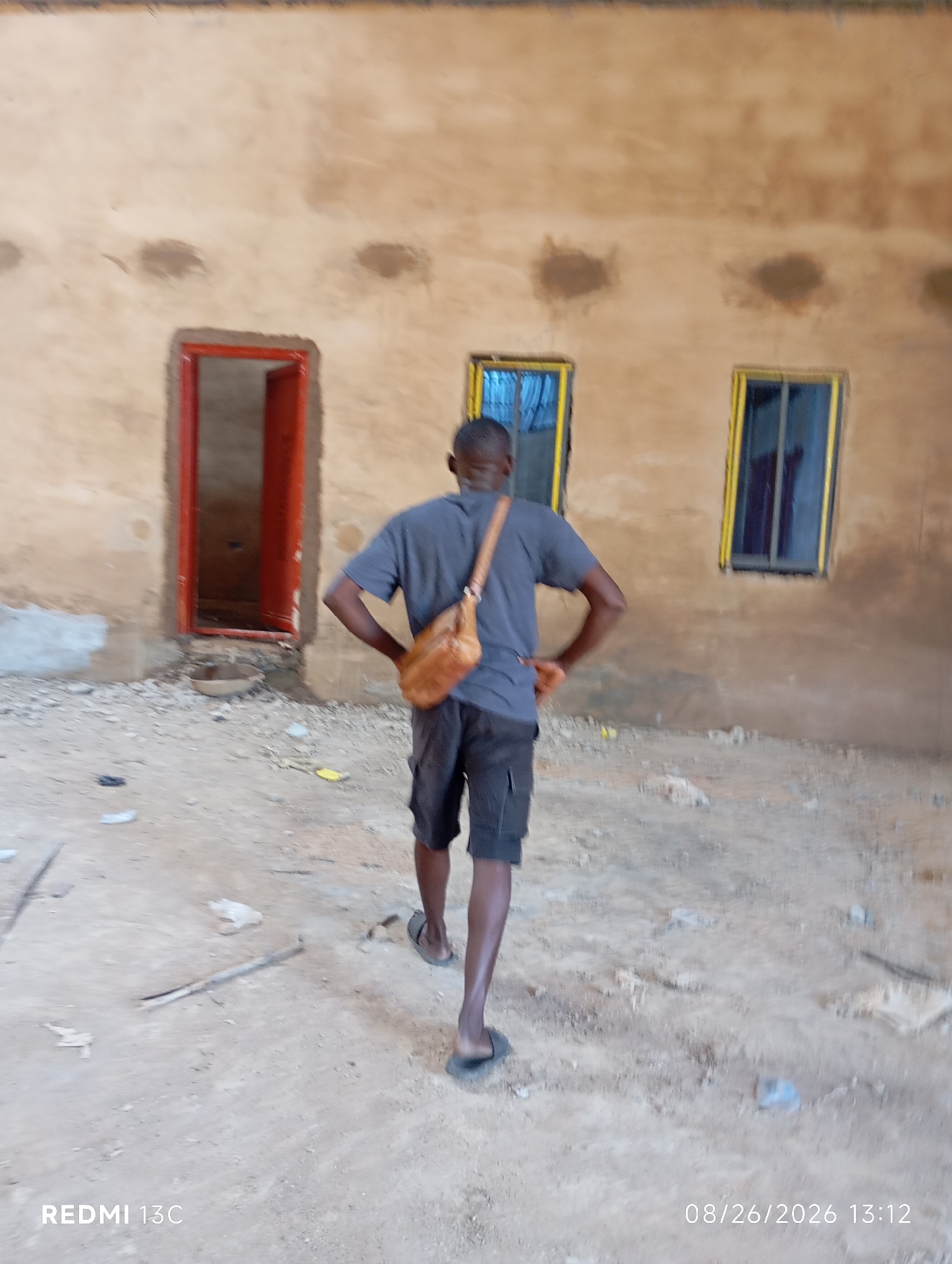 The image shows a man standing in front of a building with a neutral-toned, textured wall. There is an open doorway with a red frame to the left of the man, and two windows with yellow frames to his right. The ground is covered in dirt and debris. The man is wearing a grey t-shirt and dark shorts, with a brown satchel slung across his body. He is facing away from the camera, with his hands on his hips. The lighting suggests it is daytime. The scene appears to be an outdoor area, possibly a construction site or an undeveloped part of Shendam, Nigeria. The timestamp "08/26/2026 13:12" is visible in the bottom right corner of the image.