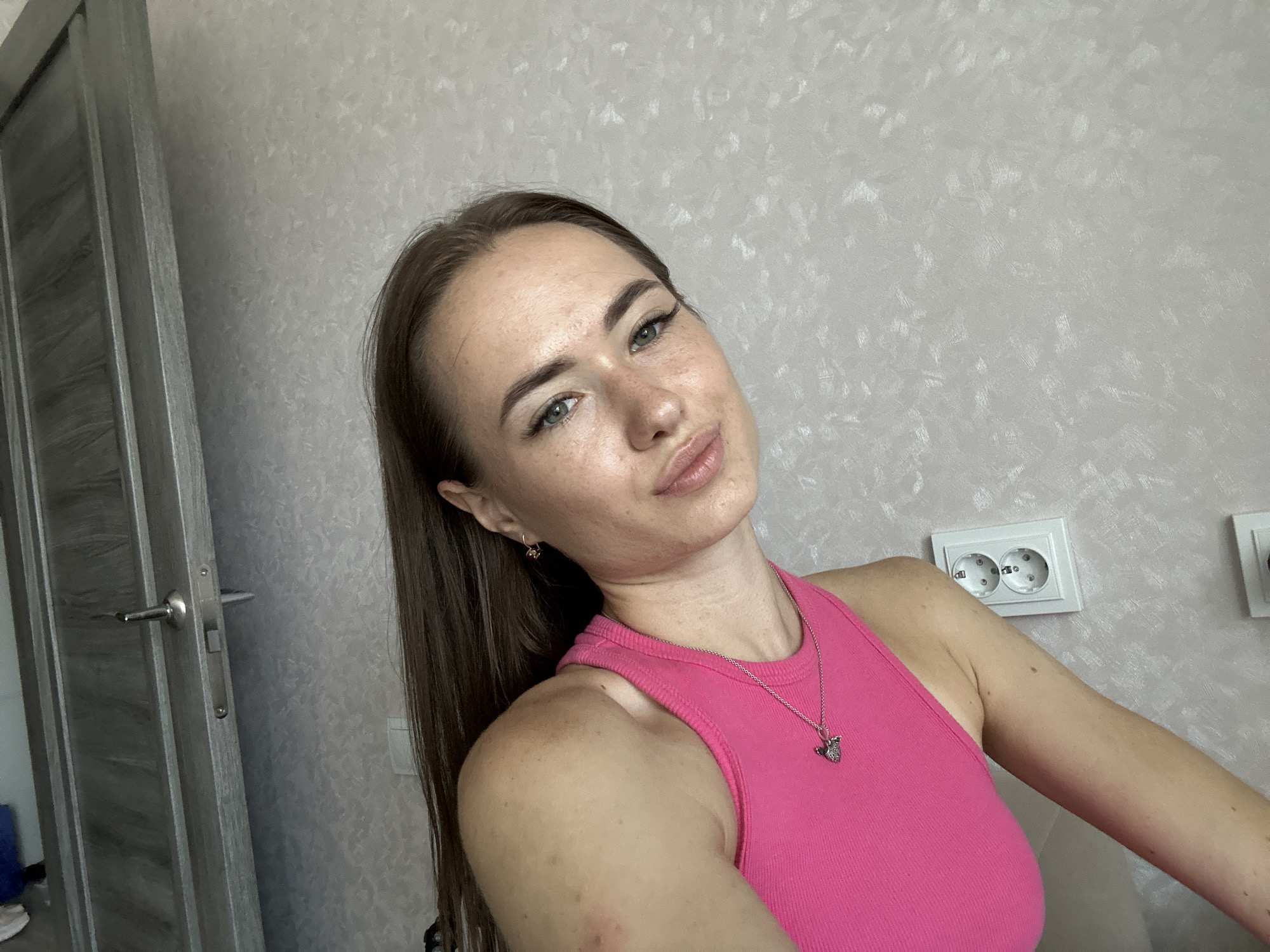 A young woman with long brown hair and green eyes is photographed indoors, likely in a bedroom or living area. She is wearing a bright pink tank top and a silver necklace with a pendant. Her head is tilted to the left, and she is looking towards the camera with a slight smile. A portion of a gray door with a silver handle is visible on the left side of the image, and a white electrical outlet is mounted on the wall behind her. The background consists of textured, light-colored wallpaper.