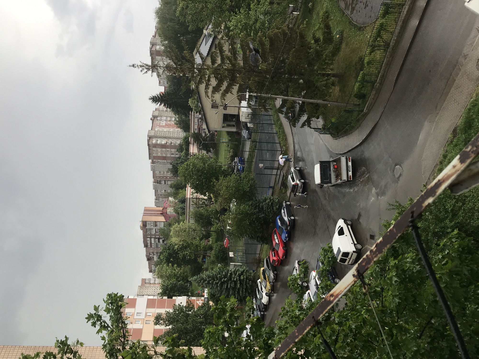 The image depicts a street scene in Ternopil, Ukraine on a cloudy, overcast day. Several multi-story apartment buildings are visible in the background. A row of cars is parked along the left side of the street including a white van parked at an angle to the curb, closer to the viewer. Further up the street, there's a work truck with its back open. Two individuals appear to be interacting near the work truck. The street is wet, suggesting recent rainfall. A metal fence and lush green trees are in the foreground, giving the impression that the photo was taken from a balcony or elevated position.