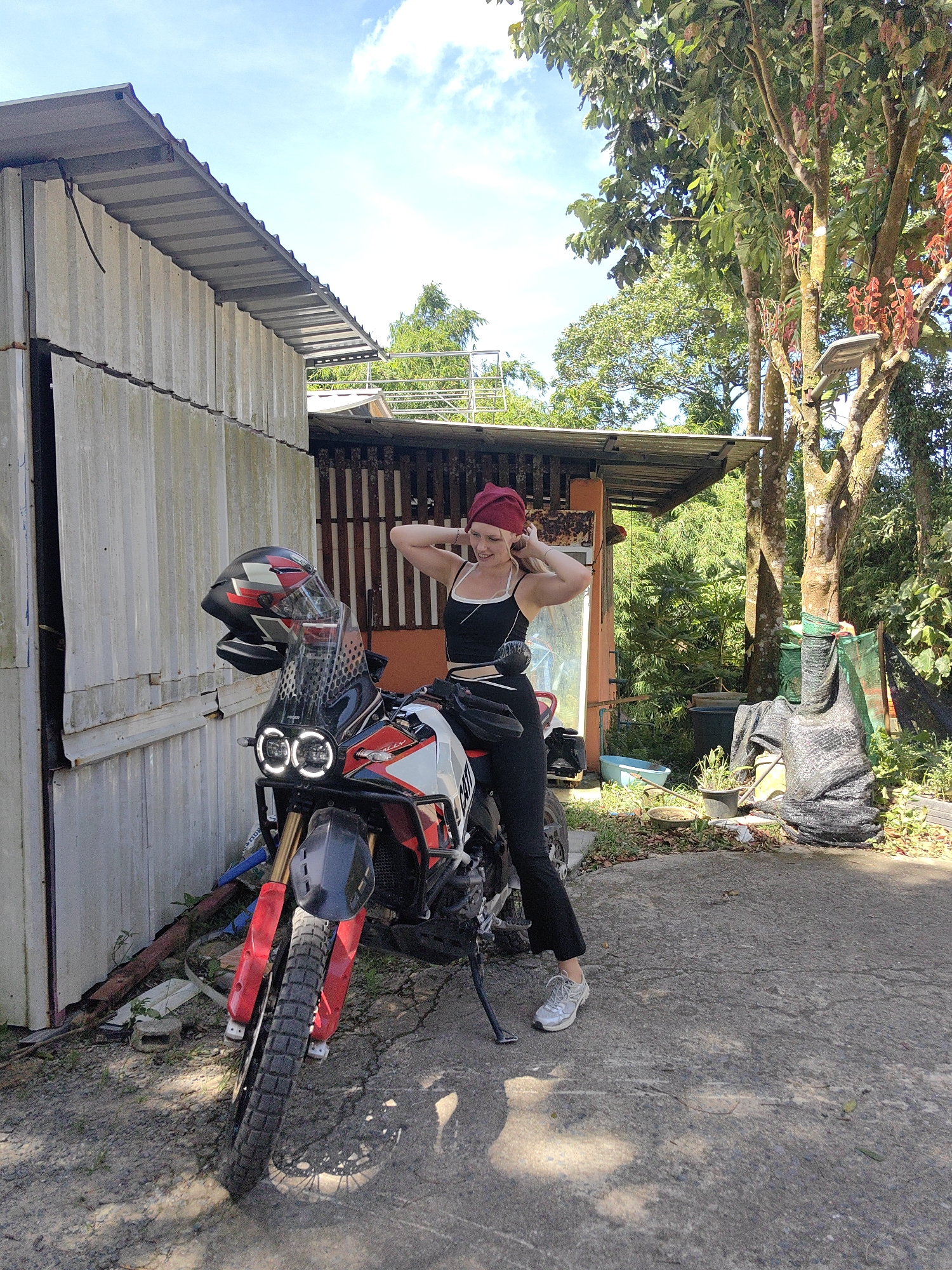A woman with blonde hair and a red headscarf is posing on a red and white dirt bike in front of a corrugated metal building. She is wearing a black tank top and black pants, and white sneakers. The woman is looking at the camera and has her hands on her head. The dirt bike has a black helmet on the handlebars and dual circular headlights. The background shows trees, foliage, and a cloudy blue sky. The scene appears to be outdoors in a rural or semi-rural area.