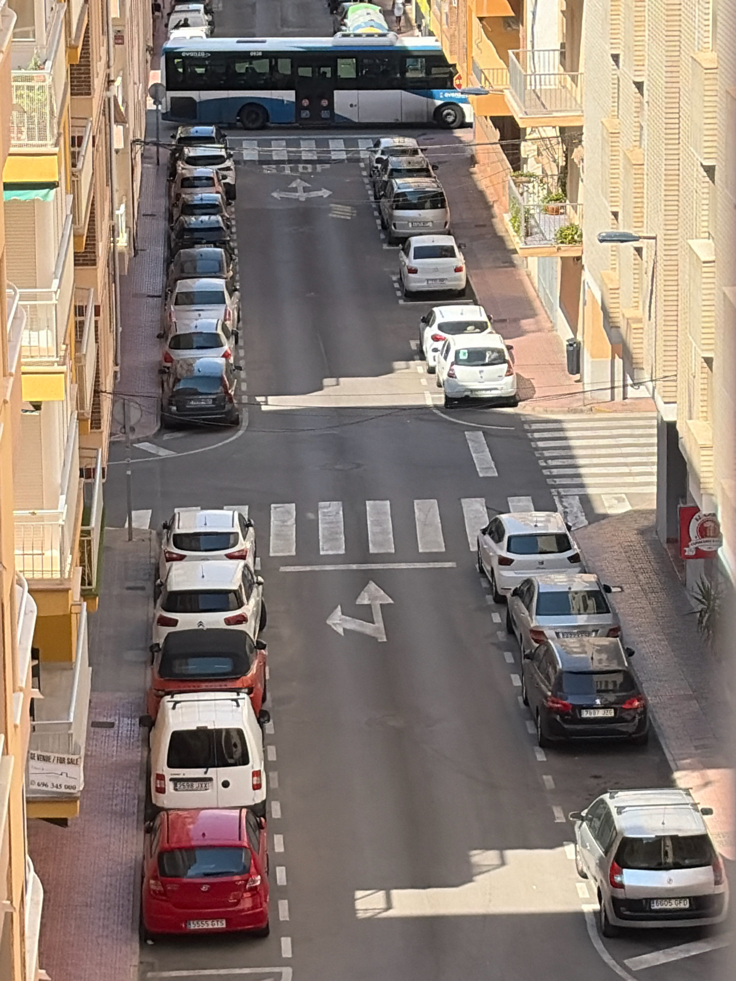The image shows a street scene in Torrevieja, Spain, likely during daylight hours. A white and blue bus is seen on the road, followed by a line of parked cars. The cars are parked on both sides of the street, which appears to be a residential area with multi-story buildings. Pedestrian crossings are visible on the road. There are no people prominently featured in the image. The environment is urban, characterized by paved roads, sidewalks, and building facades.