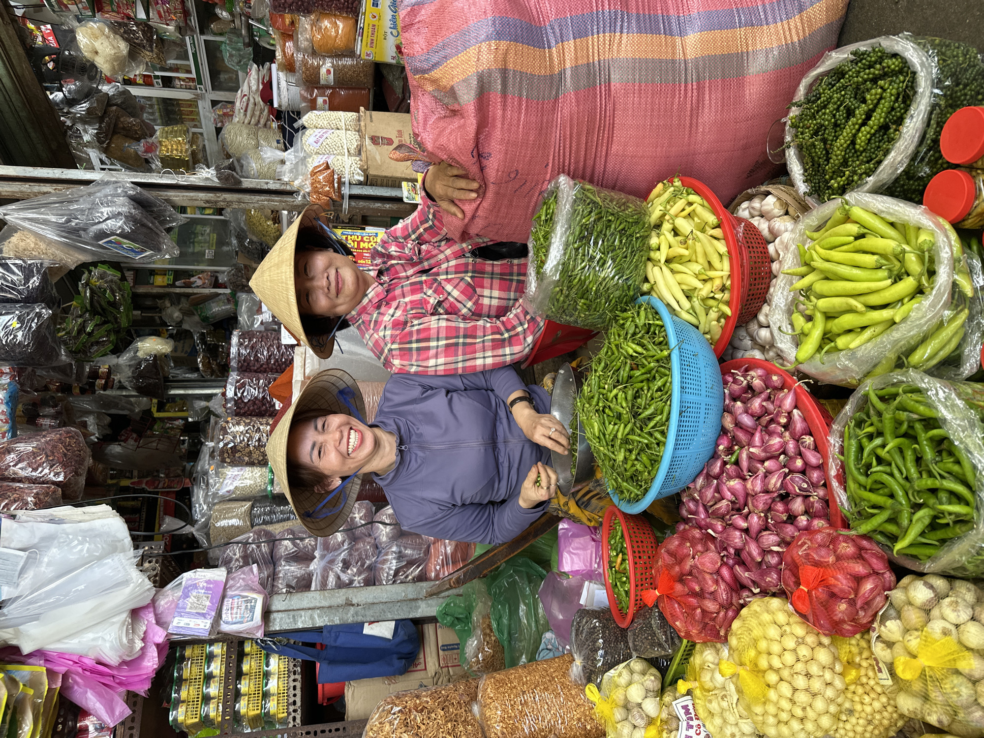 This image depicts a vibrant market stall in Đà Nẵng, Vietnam, featuring two female vendors surrounded by fresh produce and various packaged goods. The scene is brightly lit, showcasing an abundance of green chilies, shallots, garlic, and other spices.
In the foreground, two women, both wearing traditional Vietnamese conical hats (nón lá), are positioned behind a display of goods. The woman on the left, appearing younger, wears a purple long-sleeved top and a patterned skirt. She is actively sorting small green chilies into a metal bowl with both hands and displays a wide, open-mouthed smile. She has rings on her fingers and a watch on her left wrist. To her right, a second, older woman wears a red and white plaid long-sleeved shirt and has a gentle smile. Her left hand rests on a large, upright pink woven sack with vertical purple and orange stripes.
The market stall is densely packed with merchandise. In the immediate foreground and surrounding the women, there are numerous items: a blue plastic basket overflowing with small green chilies, a red plastic basket containing yellow chilies, large piles of red-purple shallots in a red basin, and net bags filled with light-colored bulbs (likely garlic). Clear plastic bags contain larger green chilies, black peppercorns, and green peppercorns on stems. Two small jars with red lids are visible at the bottom right. Behind the younger woman, two large clear plastic bags contain golden-brown dried items, possibly fried garlic or shallots.
The background reveals more of the market environment, with shelves stacked high with various packaged goods, predominantly in clear plastic bags, including dried ingredients and spices. Some items bear Vietnamese text. Overhead, numerous empty and full plastic bags hang from wires and racks. On the far left, a metal shelving unit holds yellow plastic crates containing jars or cans, and a blue fabric tote bag is hanging. The overall impression is a bustling and well-stocked market stall.