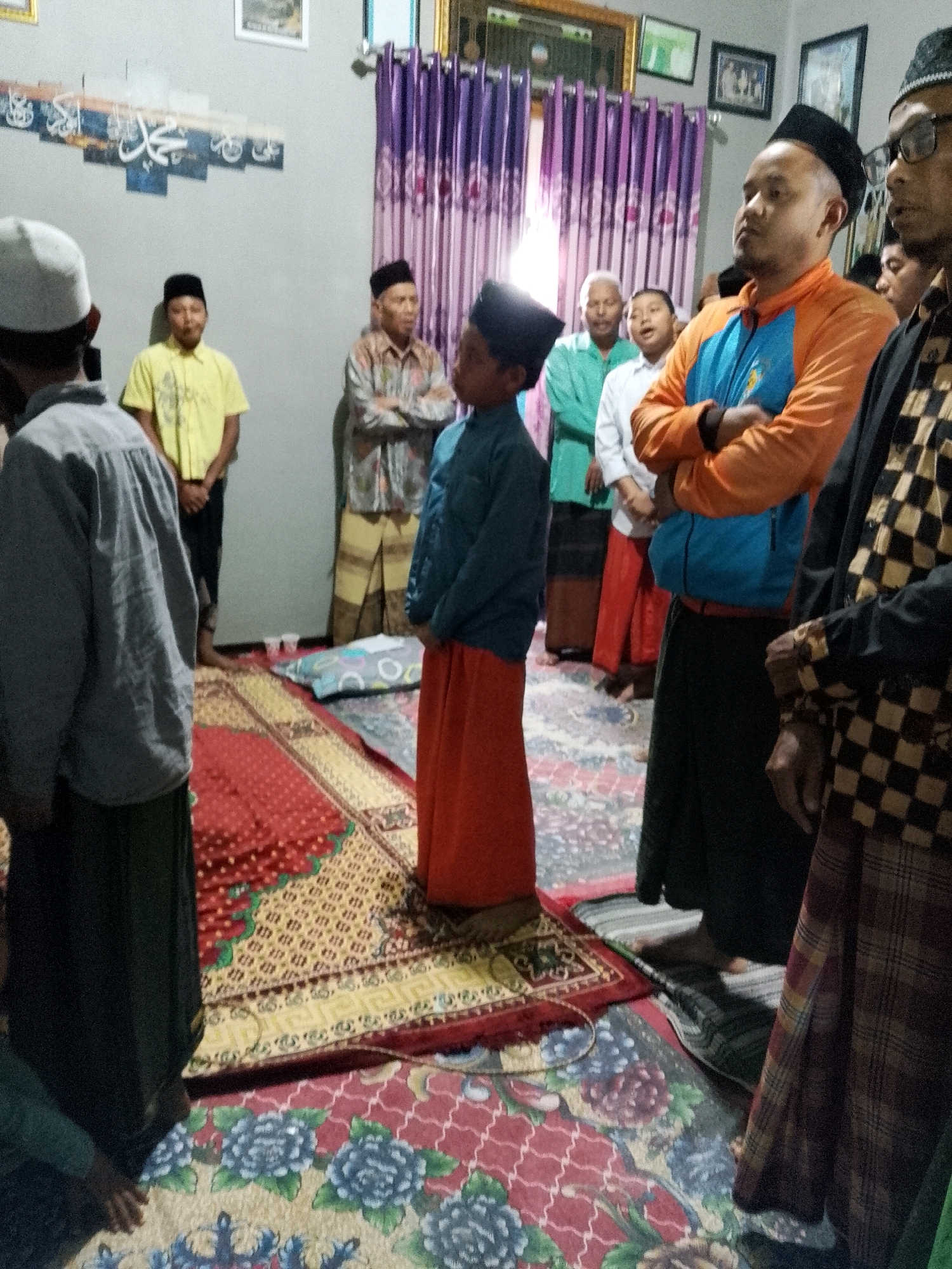 In a dimly lit room in Rejoyoso, Indonesia, a group of men and boys are gathered, possibly for a religious observance. The room is decorated with Islamic calligraphy on the wall and purple curtains. Several individuals are wearing traditional Islamic attire, including peci hats and sarongs. One boy in an orange sarong stands out, while another man in an orange and blue jacket stands with his arms crossed. The floor is covered with patterned carpets, suggesting a communal space. The overall atmosphere appears to be one of solemnity or anticipation.