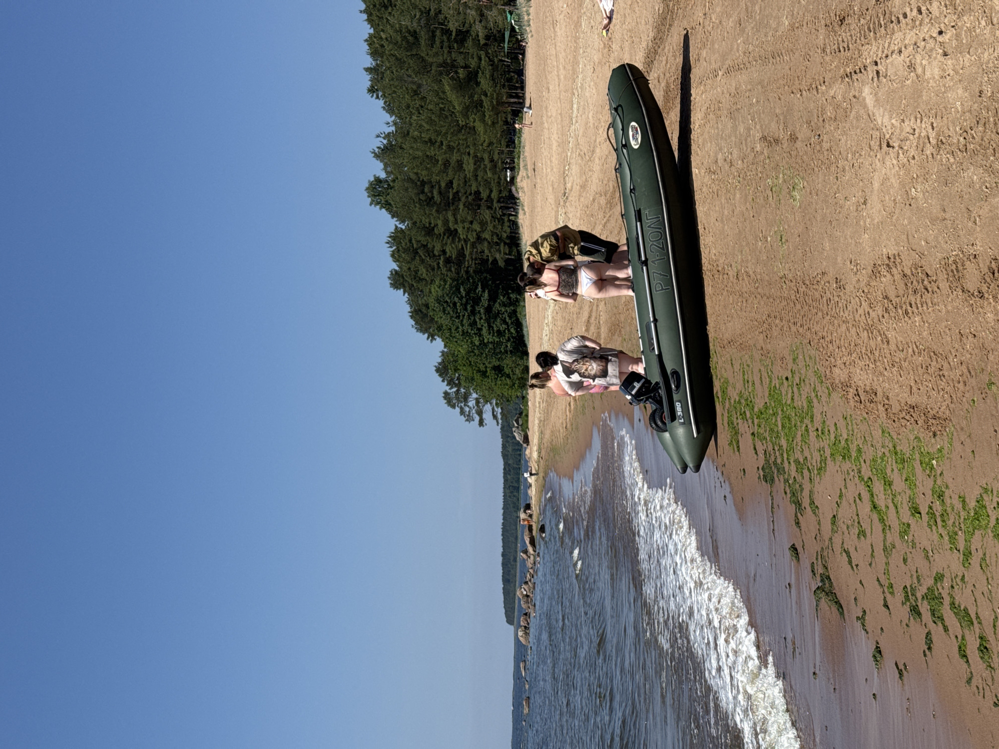 In Zelenaya Roshcha, Russia, on a sandy beach with a clear blue sky, a group of individuals stands near an inflatable boat on the shore. The boat, dark green with white lettering and an outboard motor, rests on the sand. The water, with gentle waves, meets the beach, where patches of green seaweed are visible. In the background, a dense line of pine trees borders the sandy expanse. A few other beachgoers are scattered in the distance.