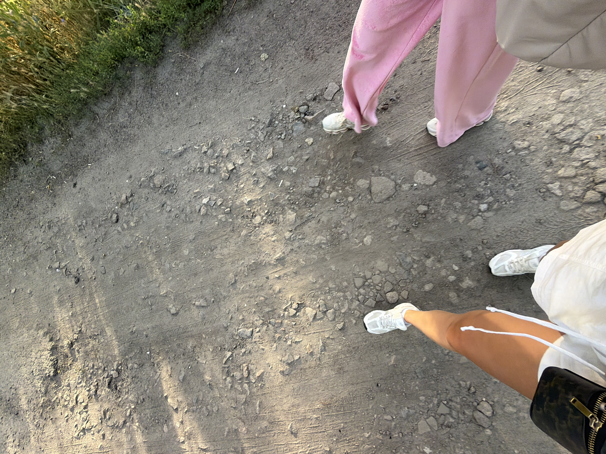 The image shows a close-up, overhead view of two people walking on a gravel path. One person is wearing light pink, wide-legged pants and white sneakers. The other person is wearing white shorts and white sneakers, with a black purse visible at the bottom right. The path is made of dirt and small stones, with some tire tracks visible. To the left of the path, there is a patch of green grass and some dry, tall grass. The lighting suggests it is late afternoon, with long shadows cast across the ground. The overall scene appears to be outdoors, possibly in a park or rural area, given the natural elements. No text is visible in the image. The image captures a casual moment of two individuals walking together.