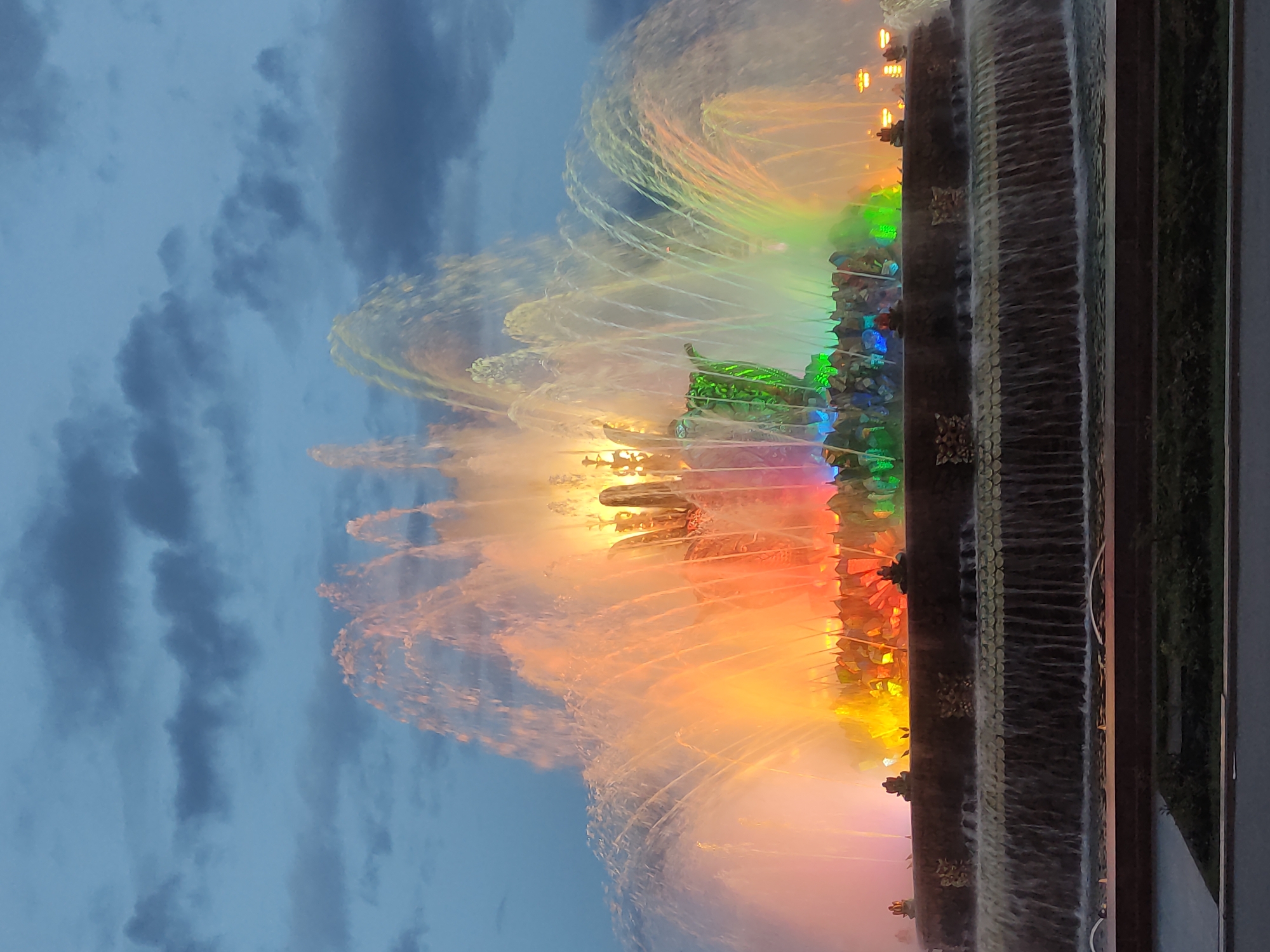 A colorful water fountain is illuminated with bright lights under a cloudy sky in Moscow, Russia. The fountain has multiple tiers and a decorative base with water cascading down the sides. The water is illuminated in various colors including red, orange, yellow, green, and blue. The sky is a mix of blue and dark gray clouds.