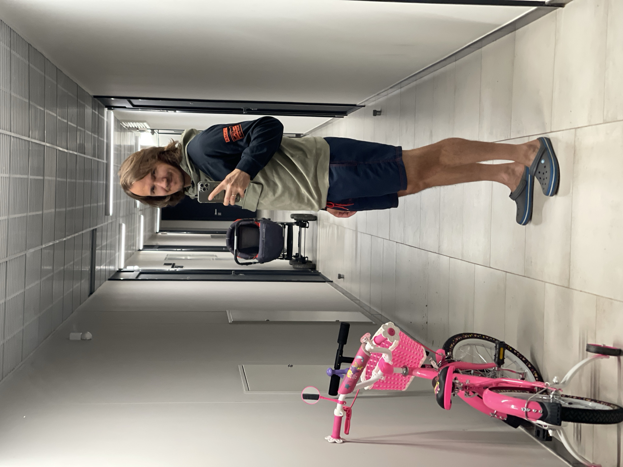 An indoor, vertically oriented image, showing a man taking a mirror selfie in a well-lit hallway. The man, with medium-length brown hair, stands centrally, looking at the camera with a slight smile. He wears a dark blue hooded sweatshirt with a light olive-green t-shirt visible underneath, dark blue shorts, and grey and blue Crocs. On his left arm, a patch on the hoodie displays orange text, partially readable as "RSBM-2". He holds a smartphone with a silver case in his right hand, reflecting in the apparent mirror.
The hallway features light-colored walls, one with a ribbed or panelled texture, and a smooth, light-toned floor. Linear lights are visible in the distance along the walls and ceiling, suggesting artificial illumination. In the background, behind the man, a dark-colored backpack or frame of a stroller is parked. To the right, a bright pink child's bicycle with training wheels and a white basket on the handlebars is visible. The bicycle has various colorful decorations, but no distinct text is legible. The overall setting suggests a residential building corridor or a similar indoor public space. The time of day is not indicated by natural light, implying artificial lighting is the primary source.