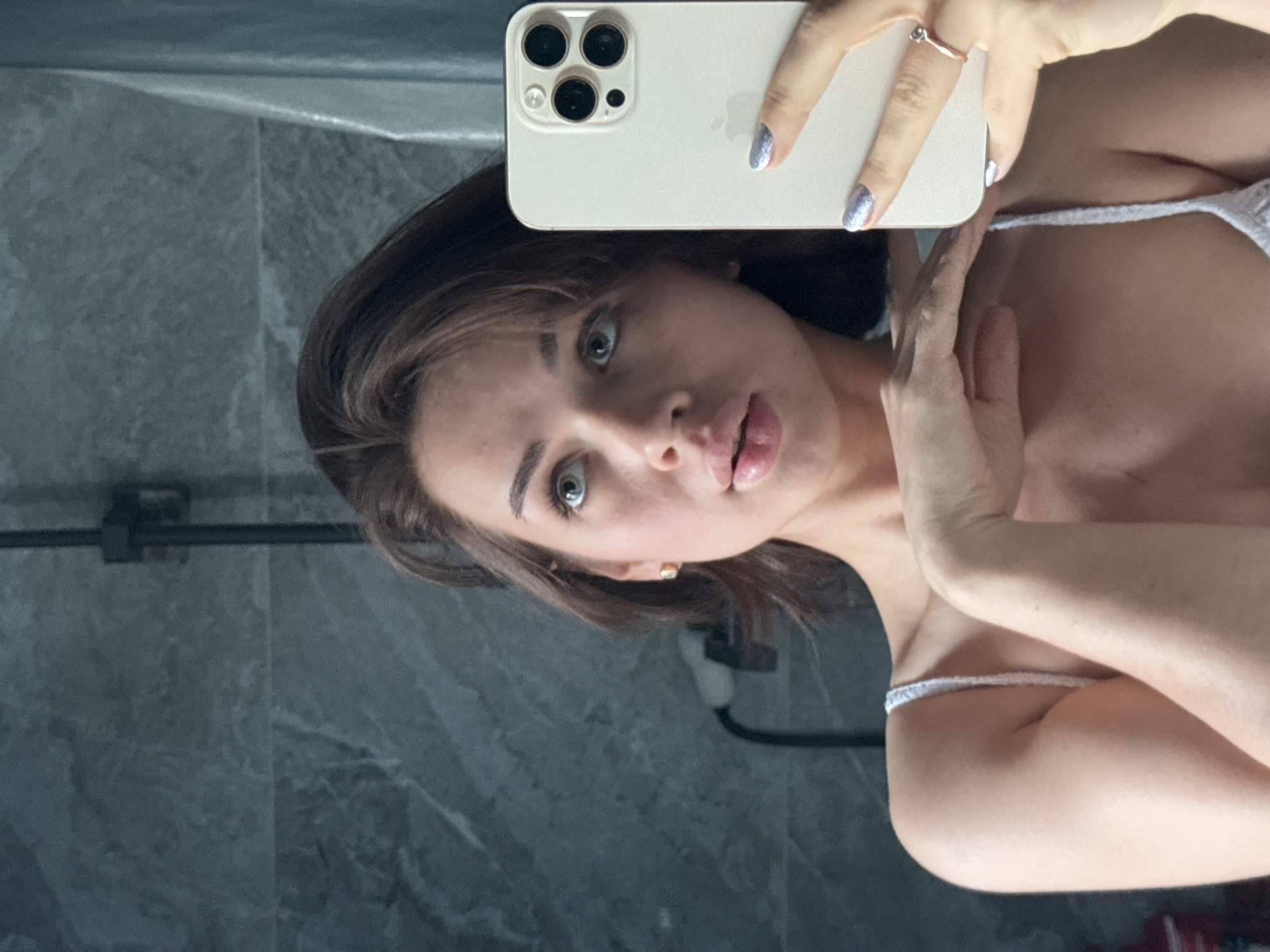A woman with brown hair and fair skin is taking a selfie in a bathroom with a grey tiled wall. She is holding a white iPhone with three camera lenses. Her fingernails are painted with silver glitter polish. She is wearing a white bra with thin straps. A black shower fixture is visible in the background. The location is not discernible from the image, so it's impossible to verify the location in the city of Лобаново, Russia.