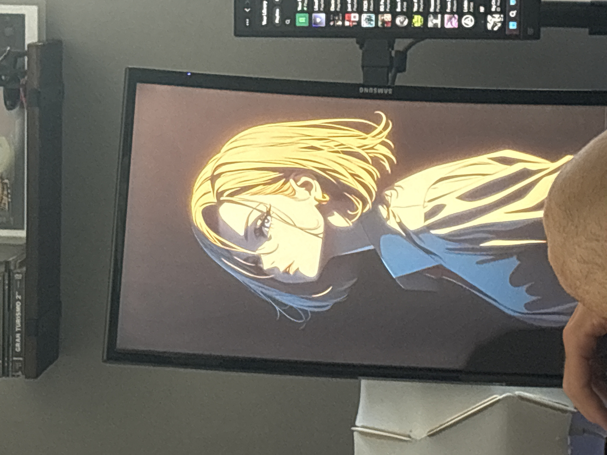 An anime-style illustration of a young woman with blonde hair, looking up and to her left, is displayed on a computer monitor. The woman is wearing a collared shirt. The monitor is positioned on a desk, with a shelf above it containing what appear to be video game cases, including one labeled "GRAN TURISMO 2". To the right of the main monitor, a secondary, smaller monitor displays a list of applications or media. Part of a person's arm and hand are visible in the lower left corner, suggesting they are interacting with the computer. The overall scene is indoors, likely a home office or gaming setup. The location of Gabrovo, Bulgaria is not evident from the image.