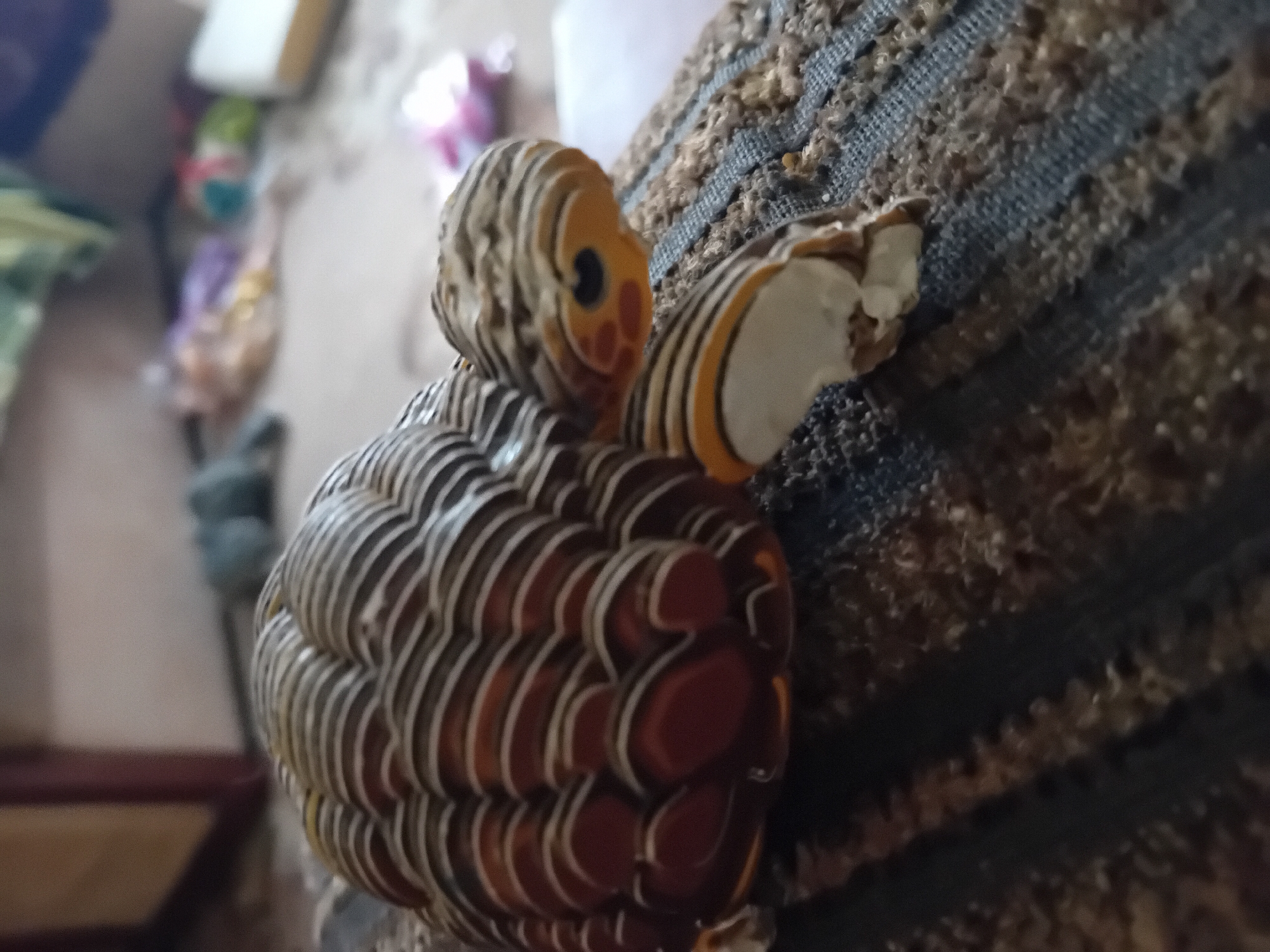 The image is a close-up, low-angle shot of a decorative ceramic turtle figurine. The turtle has a patterned shell with alternating bands of reddish-brown and white. Its head and legs are also decorated with similar striped patterns and have orange and black accents. The figurine is positioned on a textured surface, possibly a rug or upholstered furniture, with a dark blue and brown woven pattern. The background is out of focus, showing blurred shapes and colors that suggest an indoor environment with furniture and possibly other objects. The overall impression is a still life focusing on the detailed craftsmanship of the turtle figurine. There is no information in the image to determine its location in the city of Chuhuiv, Ukraine.