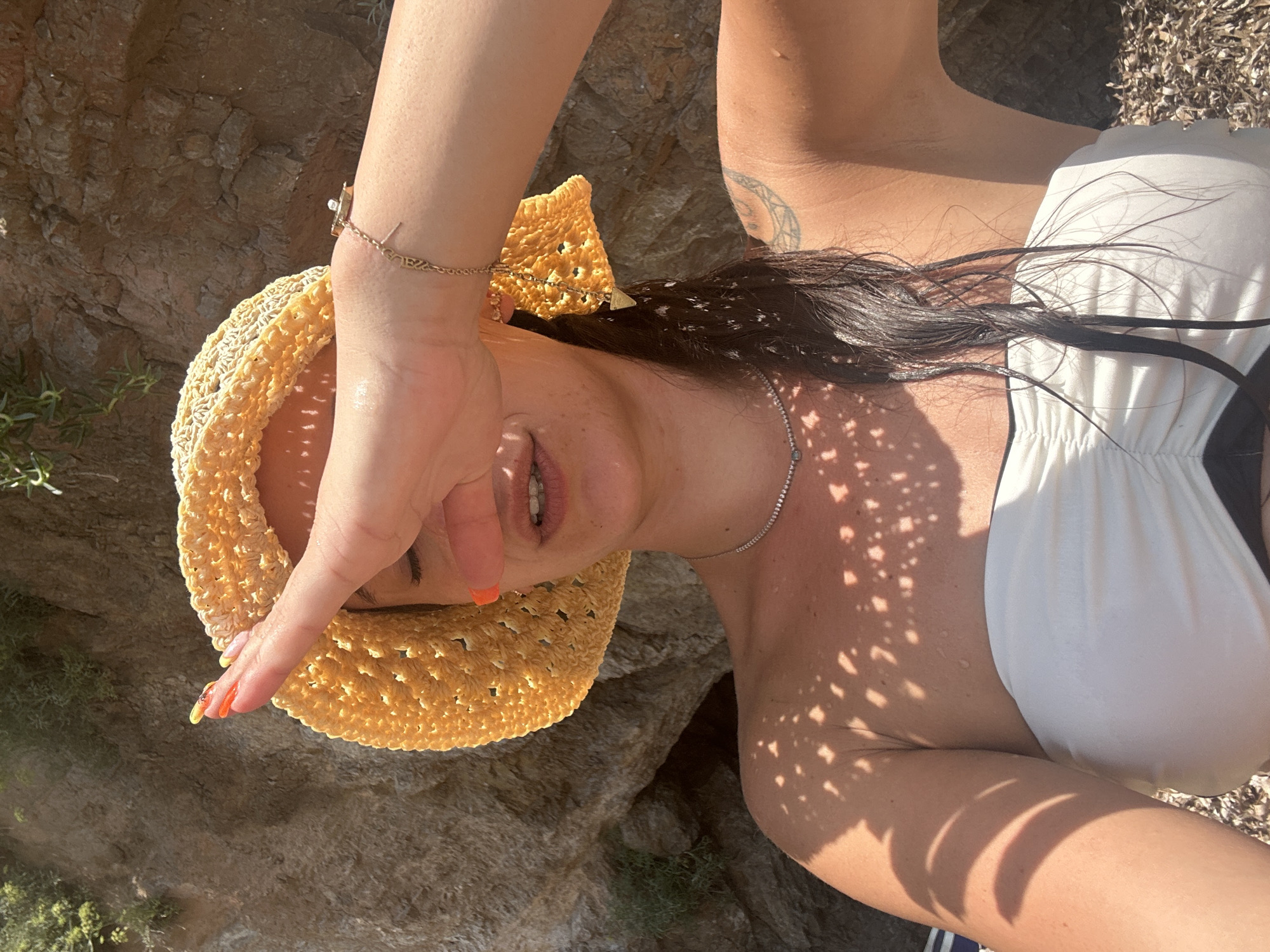A woman, wearing a yellow straw hat and a white strapless bikini top, is shielding her eyes with her right hand. Her left arm is visible, with a delicate bracelet on her wrist. She has dark, wet hair and is wearing a silver necklace. The scene appears to be a beach, with rocky cliffs in the background. The sunlight casts a patterned shadow of the hat onto her chest and shoulders. The ground is covered in pebbles and dry leaves. The location is identified as Costa Des Pins, Spain.