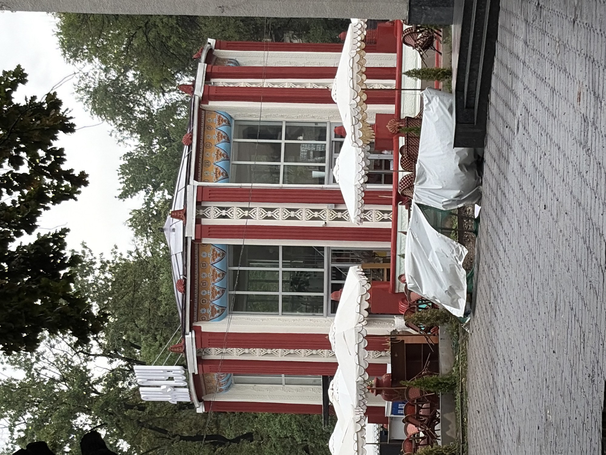 The image shows a circular building with red and white striped columns, ornate decorative elements above the windows, and a rooftop sign in a Cyrillic script. White umbrellas are set up in front of the building, and tables and chairs are visible on a patio area. Several items are covered with white tarps on the ground in front of the building. The area around the building is paved with gray cobblestones, and trees are visible in the background. A gray concrete structure with steps is to the right of the building. The scene appears to be an outdoor cafe or restaurant in Bishkek, Kyrgyzstan, with the white tarps suggesting that the establishment may be closed or preparing for inclement weather.