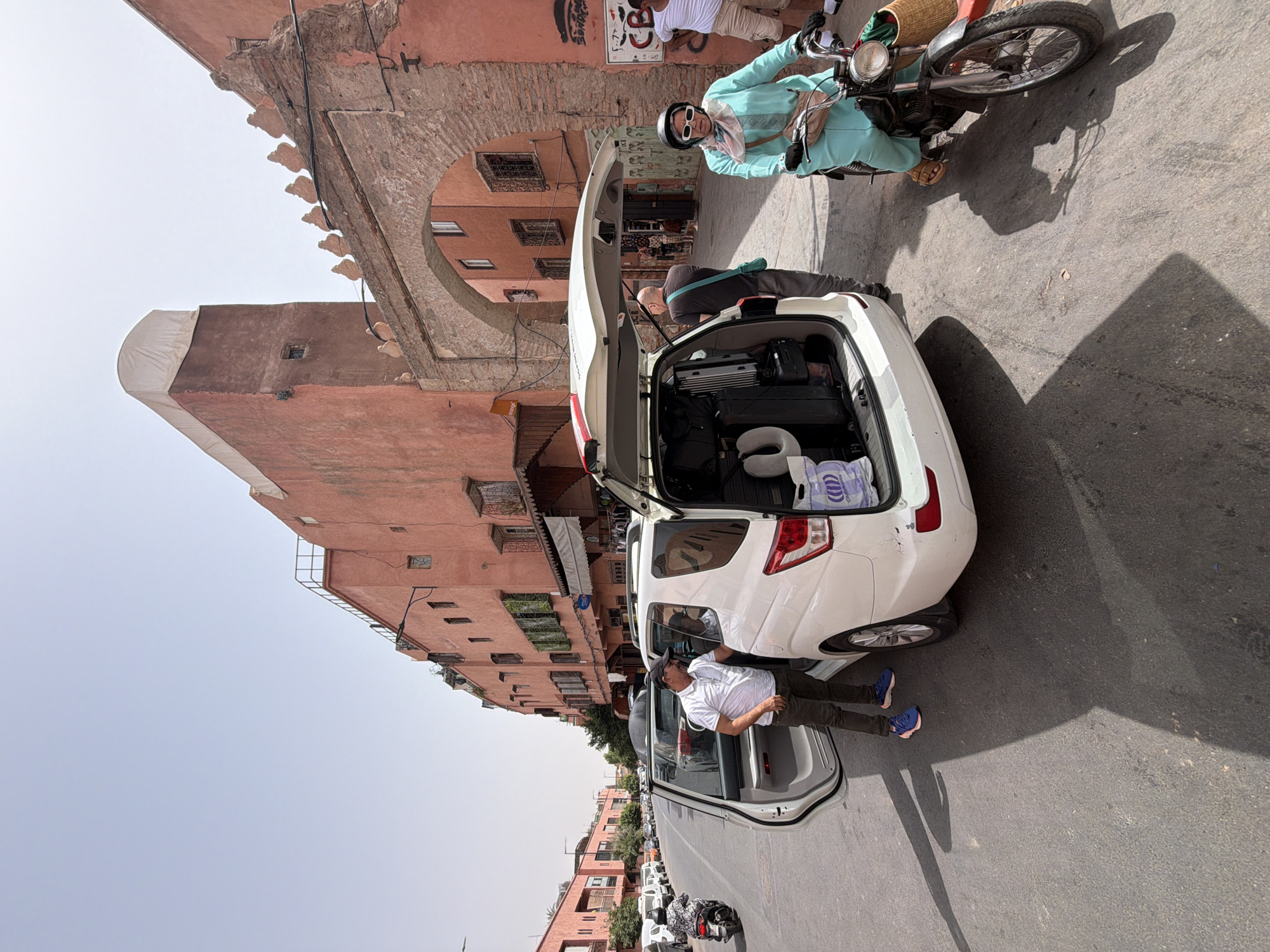 In Marrakesh, Morocco, a white minivan with its rear hatch open is parked on a narrow street with reddish-pink buildings. The minivan is loaded with several pieces of luggage, including suitcases and a neck pillow. A man in a white polo shirt and dark pants, wearing a black baseball cap, stands by the open driver's side door of the minivan, seemingly interacting with someone inside. Another man in a dark shirt and grey pants stands near the open hatch, facing away from the camera. To the right, a woman wearing a light blue traditional dress, a headscarf, sunglasses, and sandals is riding a moped. She has a woven basket attached to the front of her moped. In the background, there's an arched entryway leading into a darker alleyway, lined with shops and buildings. The sky is clear and bright blue, indicating a sunny day. The street surface is asphalt, with visible shadows cast by the vehicles and people. A few other vehicles and motorcycles are visible further down the street, and more buildings with arched windows and decorative elements line both sides of the street.