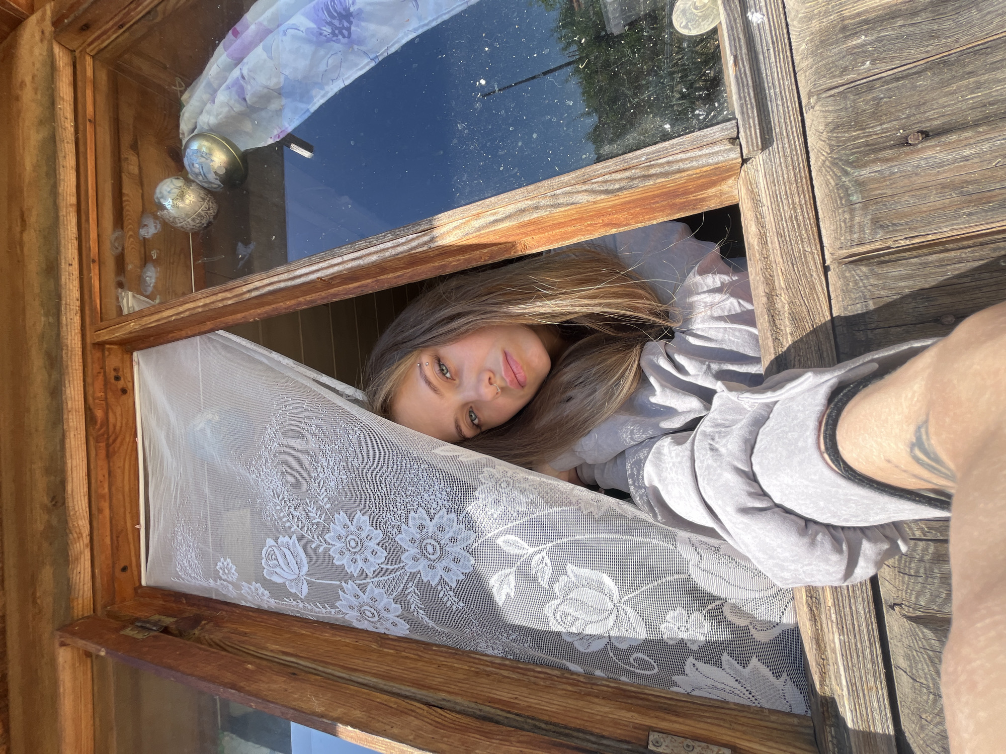 A photograph depicts a person with light brown hair and fair skin, leaning out of a wooden window. The window has a white lace curtain partially covering the opening. Outside the window, glimpses of greenery and sky are visible. The person appears to be wearing a light gray garment. There are two decorative globes on the window pane. The location is unknown.