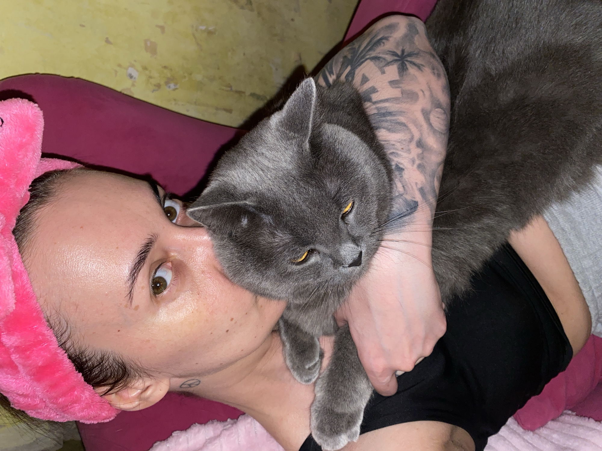 A woman with light skin is lying on a magenta surface, wearing a pink fuzzy headband. She is looking upwards with wide eyes. A grey cat is nestled against her, held by the woman's tattooed arm. The tattoo features a landscape design, including palm trees. The background appears to be a wall with peeling paint. The woman is wearing a black top. The location is not discernible from the image.