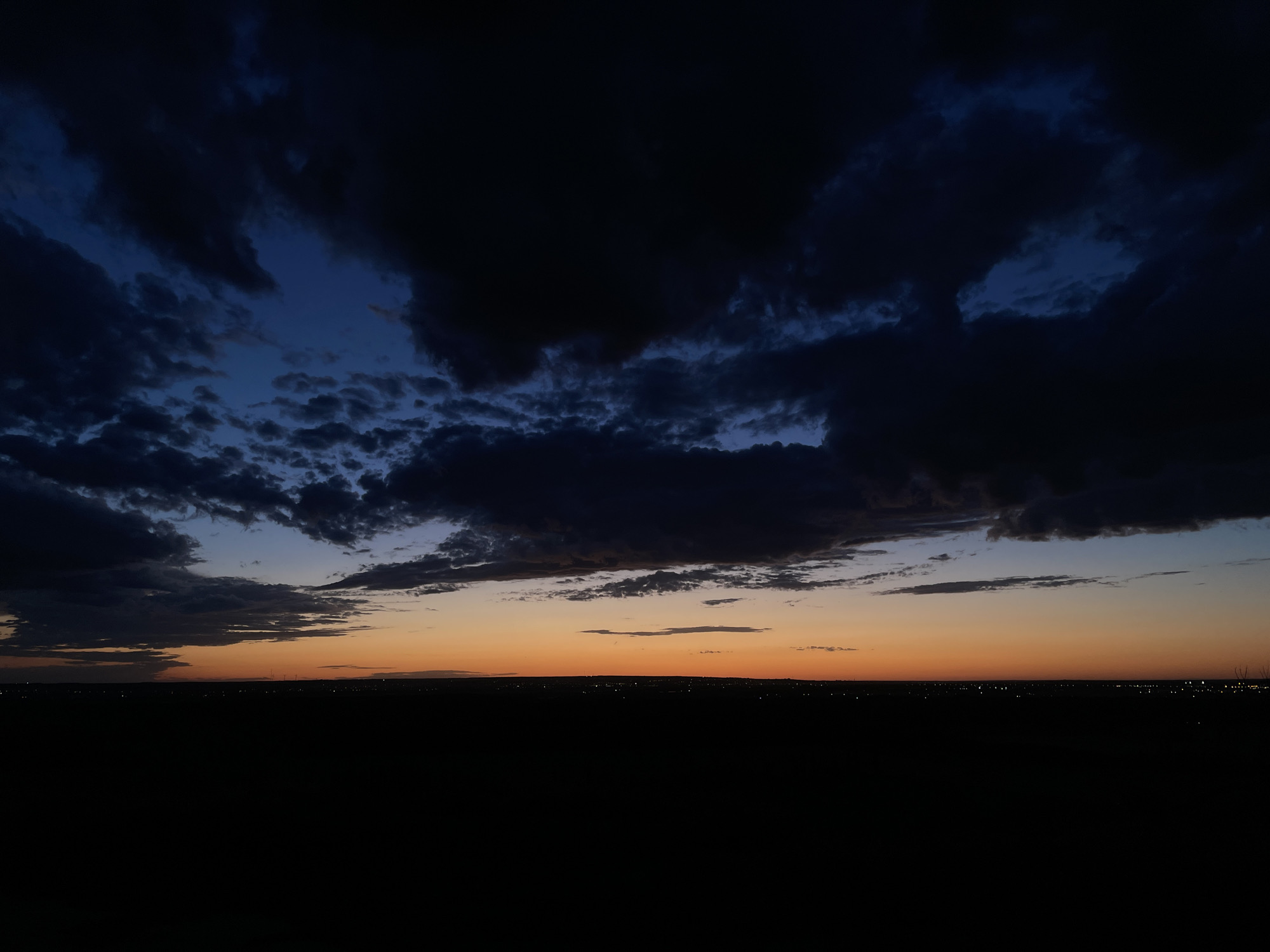 A wide shot of a night or dawn sky over Parkany, Moldova. The sky is predominantly a dark blue, transitioning to orange at the horizon. Dark clouds fill most of the frame. A distant horizon line shows faint lights, likely from buildings or streetlights. The foreground is a solid, dark mass, probably representing land or foliage. There is no sign of humans.