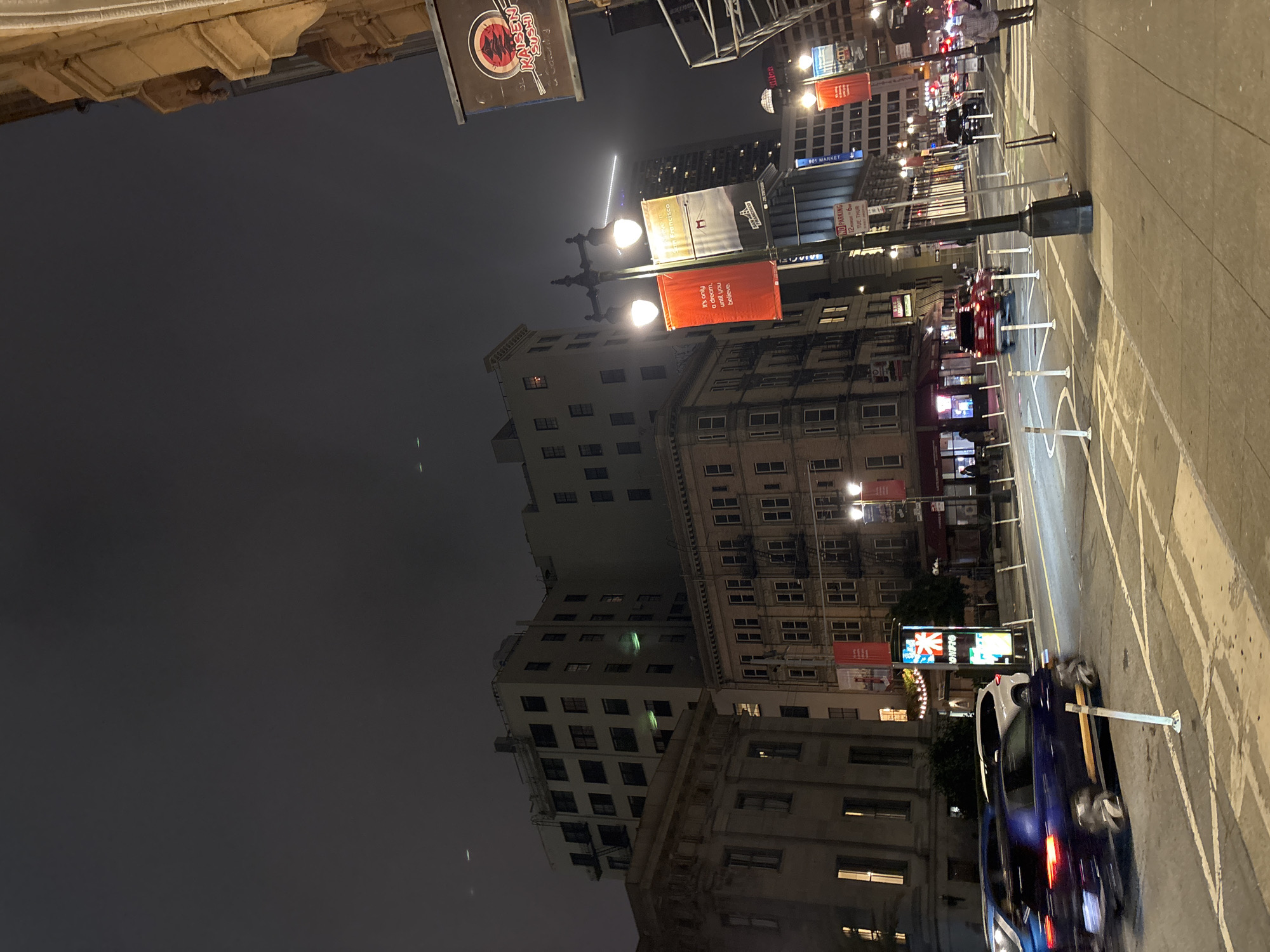 This is a night-time, street-level shot of a city intersection in San Francisco, California, United States of America. The scene is dominated by multi-story buildings with windows lit from within, indicating occupancy. The sky is dark and overcast, typical of a San Francisco evening.
In the foreground, the street is wet, suggesting recent rain or mist, and cars are visible, some in motion and blurred due to exposure time. A dark blue sedan with yellow racing stripes is driving through the intersection, its headlights and taillights illuminated. White bollards line the street, marking pedestrian walkways and traffic lanes.
The sidewalk is made of concrete and shows signs of wear. A person in a plaid shirt is walking away from the camera on the sidewalk. Several street lamps with illuminated globes line the street, providing a warm light.
Various signs and banners are visible on the buildings and poles. A sign for "KAISEN SUSHI" is prominently displayed on the right side of the image, featuring an image of a red pagoda. Red and white banners hang from lampposts, one with text that reads "It's only a dream, until you believe." and another with "SAN FRANCISCO". A "NO PARKING" sign is also visible, indicating parking restrictions.
The architectural styles of the buildings vary, from older, ornate structures to more modern high-rises. The overall impression is that of a busy urban environment at night, with the typical elements of city life – traffic, lighting, signage, and pedestrians – present.