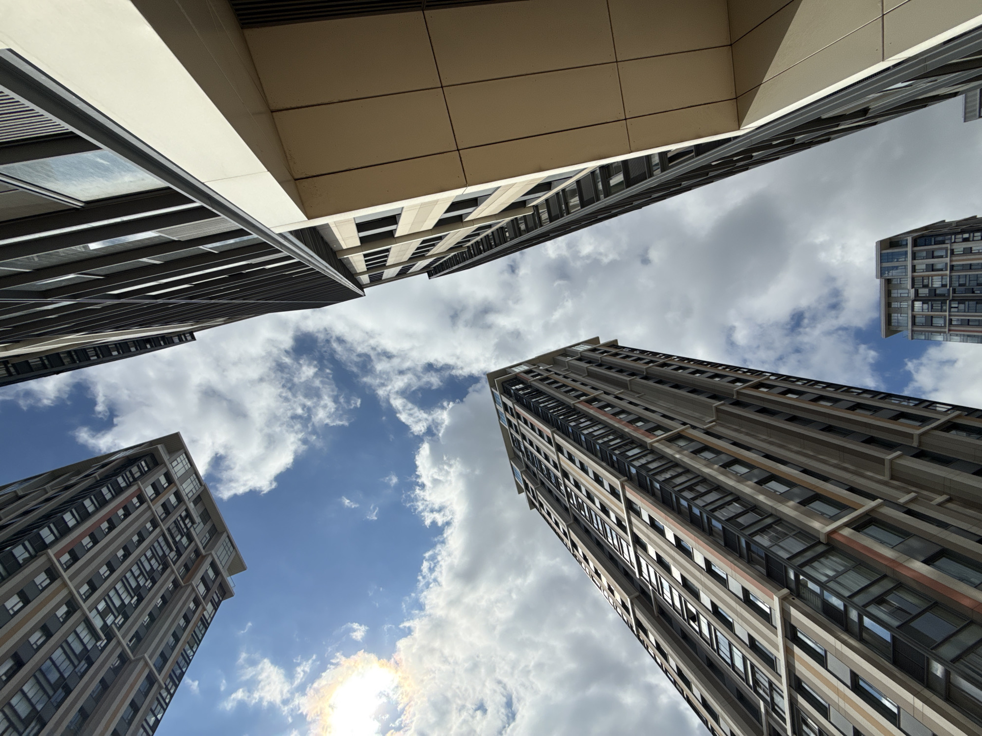 An upward-looking, low-angle shot captures a cluster of modern high-rise buildings against a partially cloudy blue sky. The sun is visible in the upper left corner, casting a bright glare. The buildings are characterized by their geometric designs, featuring numerous windows and contrasting facade materials, including beige panels and dark grey or black accents. One building on the right has a prominent beige overhang. In the lower right, another smaller building is partially visible. The image appears to be taken from street level, looking directly up at the architecture. The scene depicts a contemporary urban environment, likely a residential or business district in Moscow, Russia, given the architectural style. There are no people or vehicles visible in the frame, focusing solely on the man-made structures and the sky.
