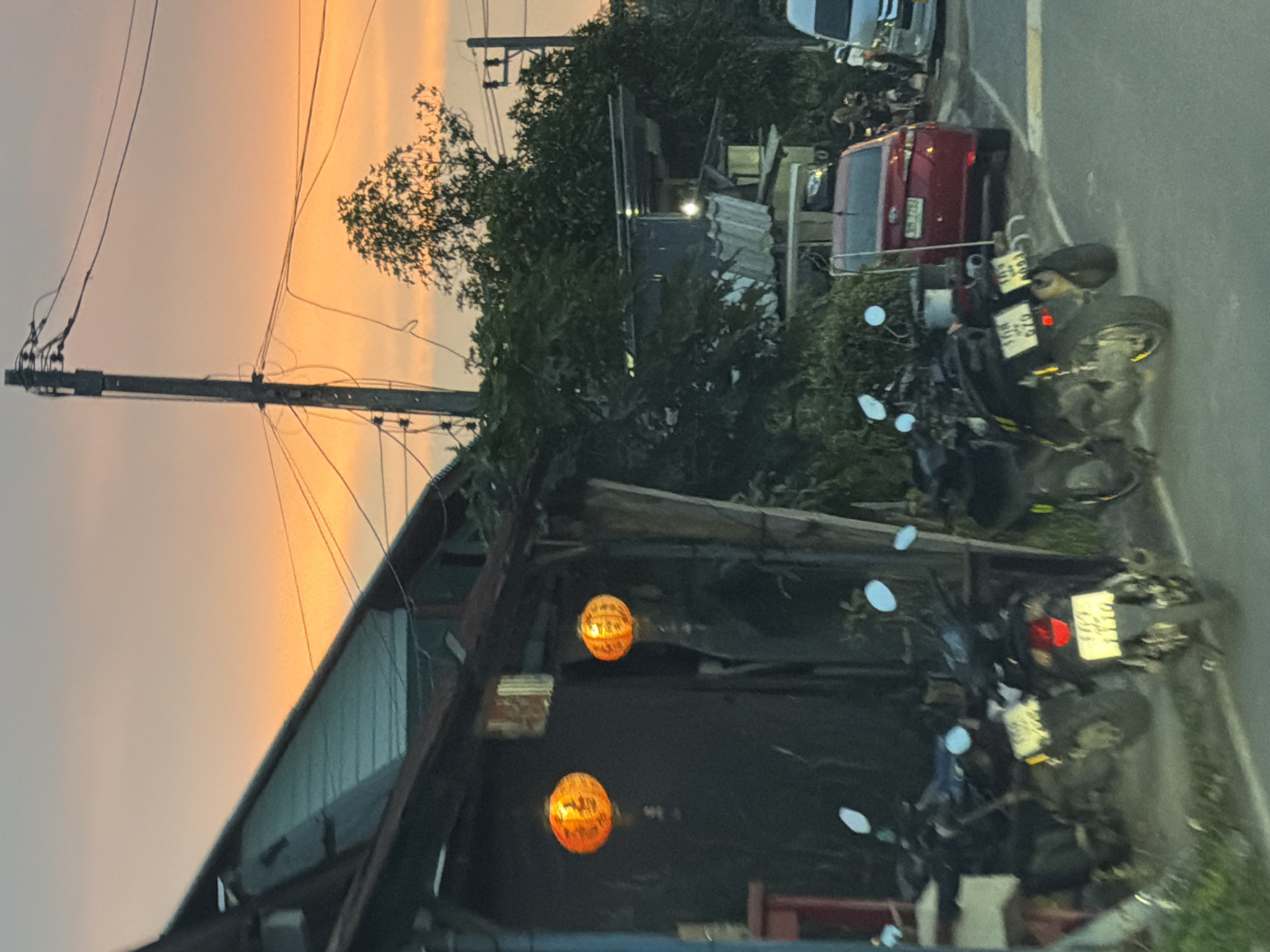 The image depicts a street scene in Choeng Thale, Thailand, during dusk. In the foreground, several motorcycles are parked along a weathered wooden structure. Two of the motorcycles, closer to the viewer, have visible license plates. The wooden structure has two illuminated orange lanterns hanging from it. Behind the motorcycles and structure, trees and other buildings are visible. A red car is parked on the right side of the street, with a white van further down. The sky is filled with the warm colors of sunset, with power lines and utility poles crisscrossing the frame. A few indistinct figures are visible in the background near the parked vehicles.