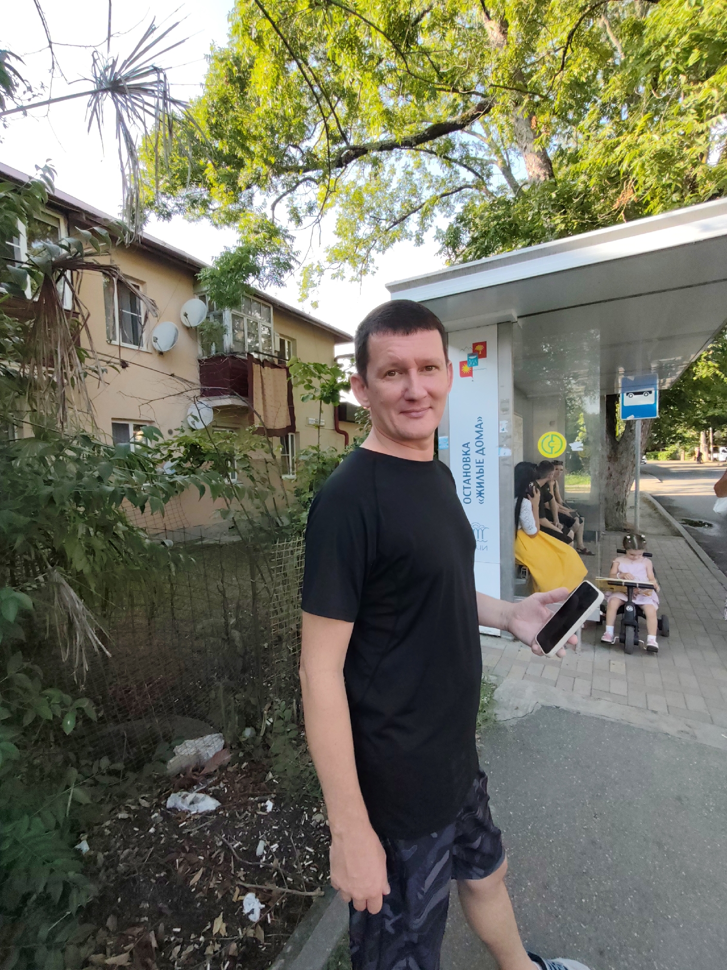 In Sochi, Russia, a man stands at a bus stop, holding a smartphone. He is wearing a black t-shirt and patterned shorts. Behind him, a bus stop shelter with informational signage is visible. Several people are seated inside the shelter, including a woman in a yellow dress and two children, one on a small ride-on toy. The background includes a multi-story building and lush green trees. The ground in front of the building appears somewhat untidy with debris.