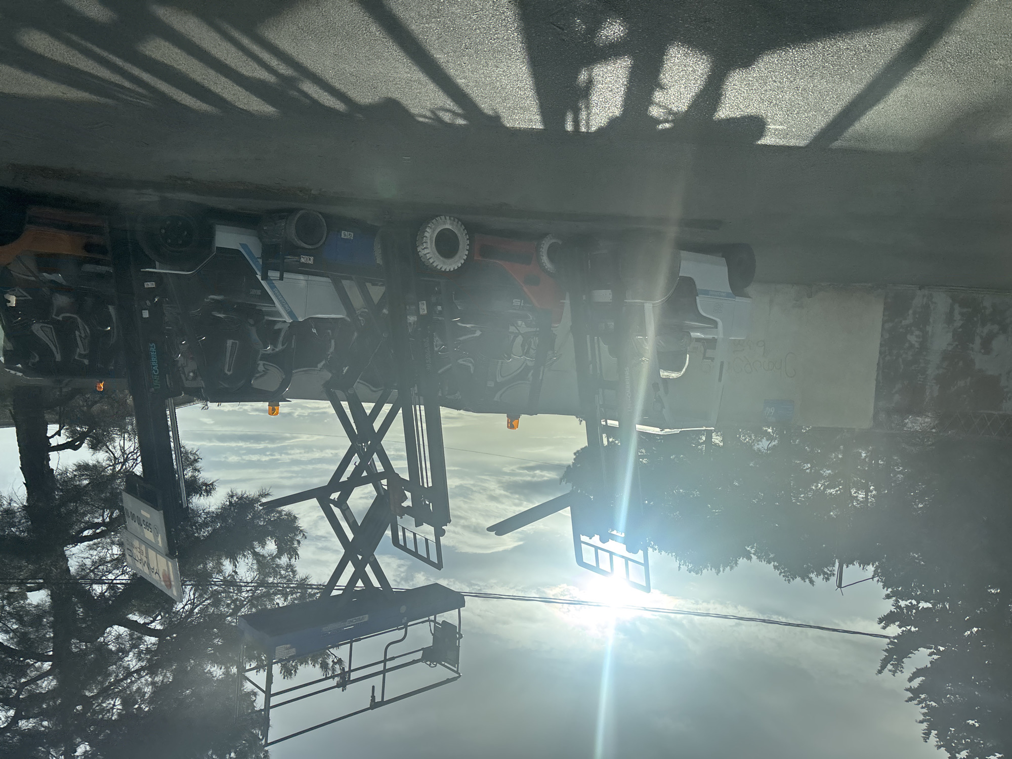 The image depicts the underside of a vehicle transporting ski lifts, likely for installation or maintenance, in Tbilisi, Georgia. The vehicle is parked on a paved surface casting tree branch shadows. There is writing on a concrete wall partially visible behind the vehicle. A tree is also visible near a ski lift in the foreground. Sunlight is present in the scene.