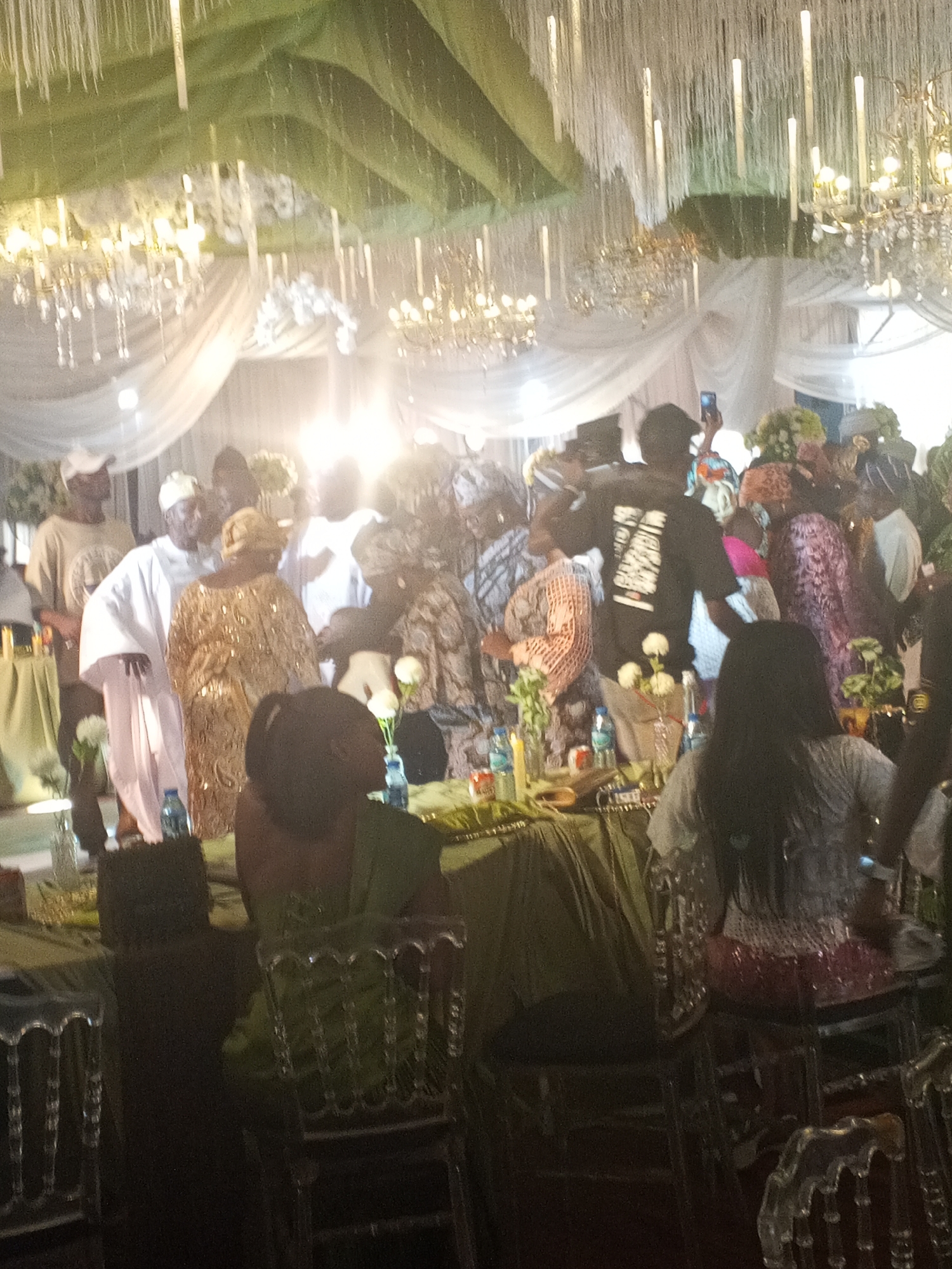 The image shows an indoor gathering of people, likely a celebratory event like a wedding, held in Barakin Lerawi, Nigeria. Several people are standing, and some are seated at tables covered with green tablecloths. There are clear, decorative chairs at the tables. Decorations include chandeliers, white flowers in vases, and ceiling draperies. One person is holding a cell phone up, seemingly taking a photo or video. Clothing styles indicate a formal or semi-formal occasion with traditional elements.
