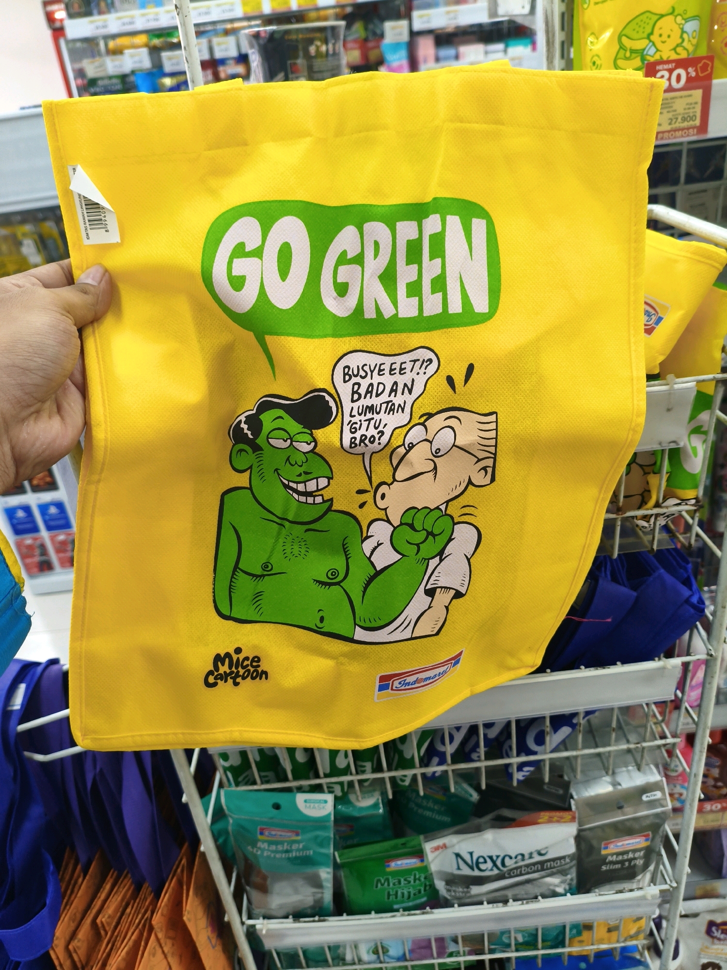 A bright yellow tote bag with "GO GREEN" printed in large green letters is held by a person's left hand. The bag features a cartoon illustration of two characters: a green, muscular figure with a wide grin and a bald, bespectacled figure wearing a white shirt and also smiling. The green character has text above its head that reads "BUSYEEET!? BADAN LUMUTAN GITU, BRO?". Below the cartoon, the words "Mice Cartoon" are written in a stylized font, and an "Indomaret" logo is present. The bag is displayed on a metal rack in what appears to be a retail store, with shelves of products visible in the background. The racks also hold other items, including blue and purple bags, and several packages of face masks. The setting is indoors, likely a convenience store or supermarket.