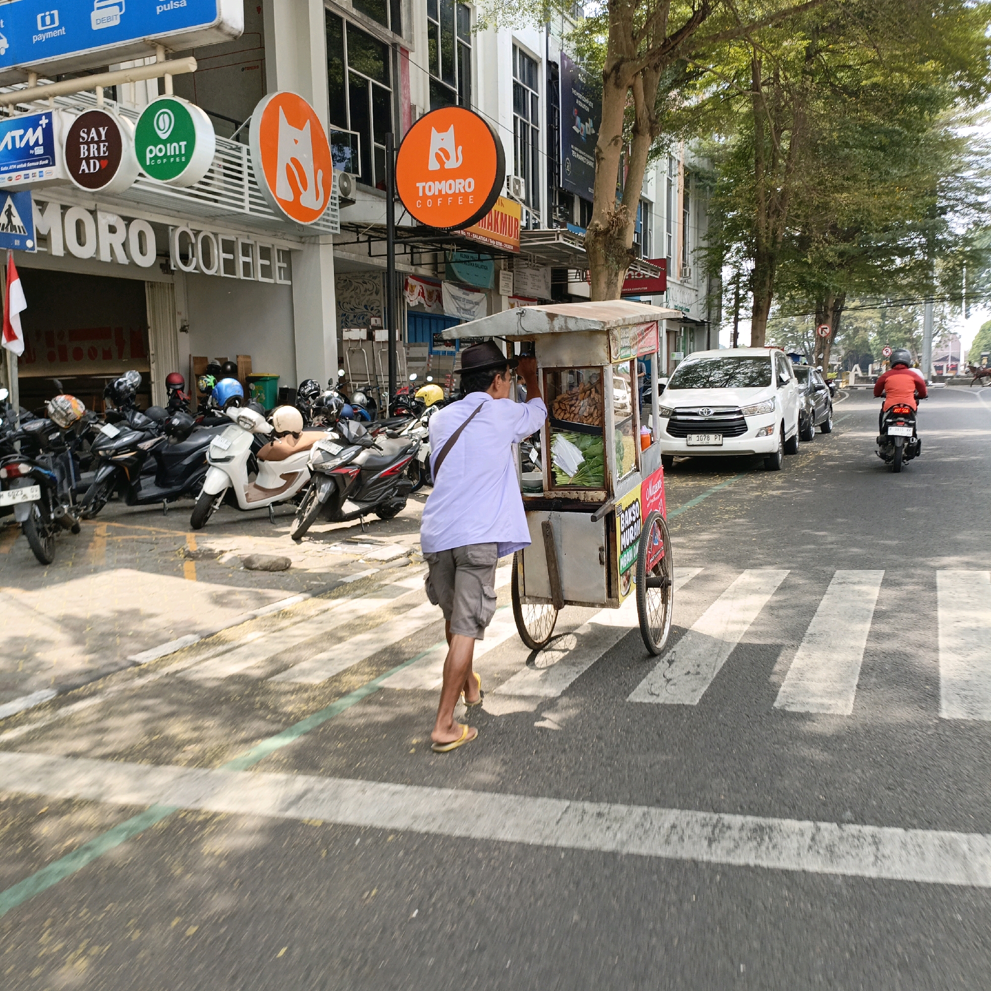 The image depicts a street scene in Salatiga, Indonesia, likely on a bright, sunny day with dappled sunlight filtering through trees. In the foreground, a man wearing a brown hat, a light purple long-sleeved shirt, and gray cargo shorts pushes a food cart across a pedestrian crossing. The cart has a canopy and displays various food items, including what appear to be fried snacks and wrapped green vegetables, with signage in Indonesian that includes "BAKSO MURAH." A brown strap crosses his left shoulder. The cart has two wheels on the right side, one of which is partially visible.
The street is paved with asphalt and marked with white pedestrian crossing stripes and a thin green line. To the left of the pedestrian crossing, a row of parked motorcycles is visible, some with helmets on them. Behind the motorcycles is the facade of a building with signage including "MORO COFFEE," "ATM," "SAY BREAD," and "POINT COFFEE." Circular signs with logos for a cat and coffee are also present.
Further down the street, a white SUV (Toyota Innova) is parked, and a black car is behind it. A person on a red motorcycle is riding away from the camera on the right side of the street. The environment is urban, with buildings lining the street and trees providing shade. The overall impression is a bustling street scene in a commercial area of Salatiga.