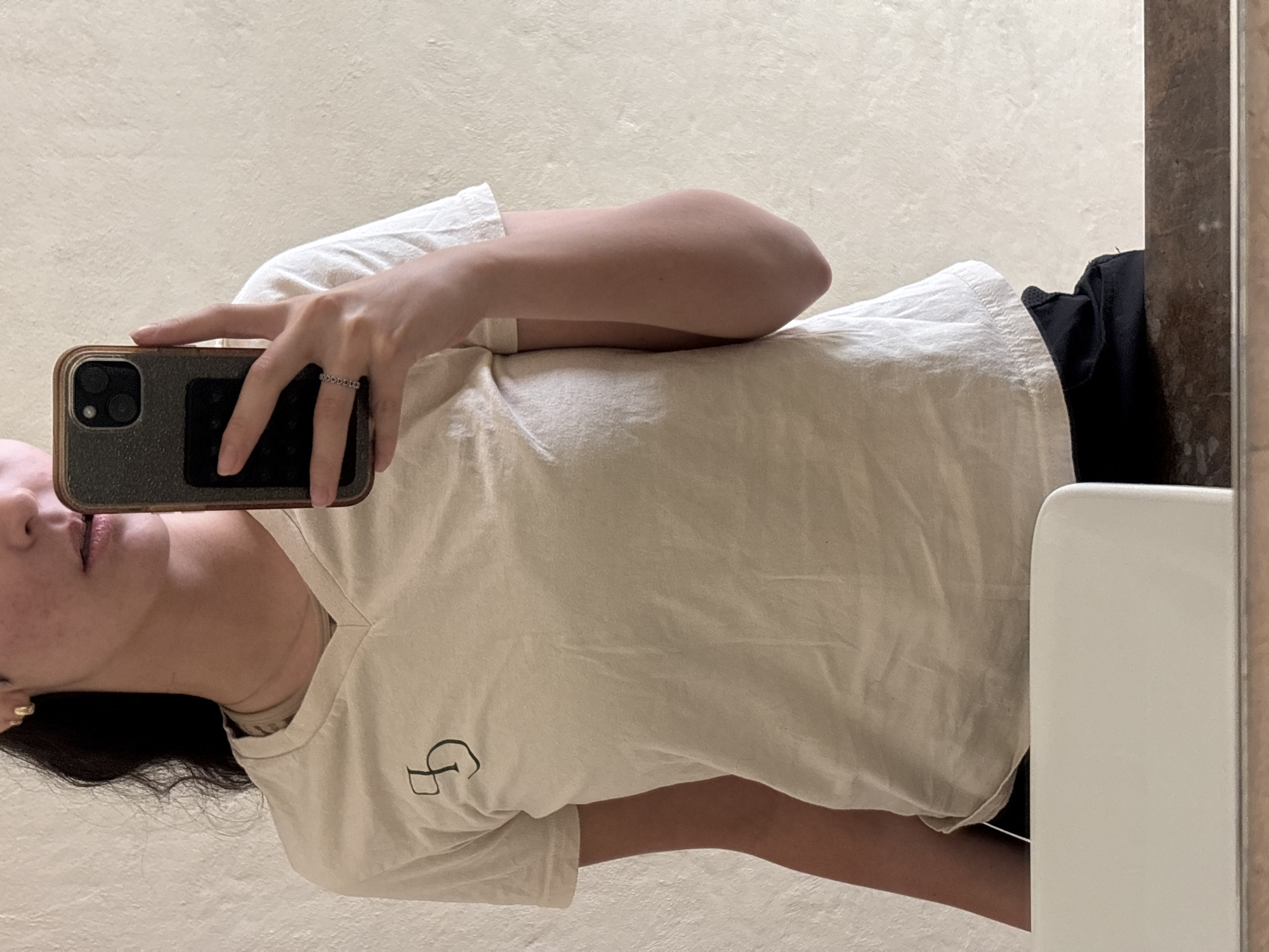 A person is taking a selfie in what appears to be a bathroom. The person is wearing a light-colored T-shirt with a graphic on the front. They are holding a smartphone in their left hand. Parts of their face, including lips, nose, and parts of dark hair are visible. The background consists of a textured wall and a mirror. There is no evidence to suggest the image was taken in San Bernardino Tlaxcalancingo, Mexico.