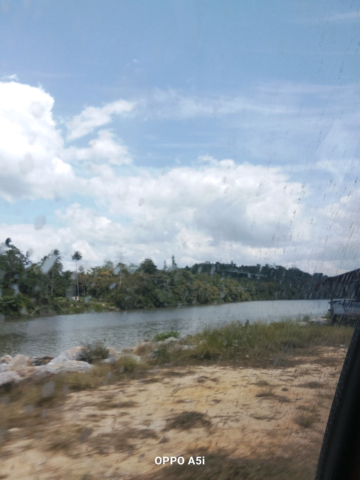 A serene landscape shot captures a wide river flowing through a lush green forest, under a partly cloudy sky. The foreground is a dirt path with dry grass. The water reflects the sky and the dense foliage along the riverbank. Several palm trees are visible among the other trees. On the right side of the river, there's a hint of a structure, possibly a building or boat dock. The image seems to be taken from a moving vehicle, as indicated by the slight blur and water droplets on the window. The location is identified as Puusangi, Indonesia. The bottom right corner of the image displays "OPPO A5i" which is likely the device used to capture the photograph.