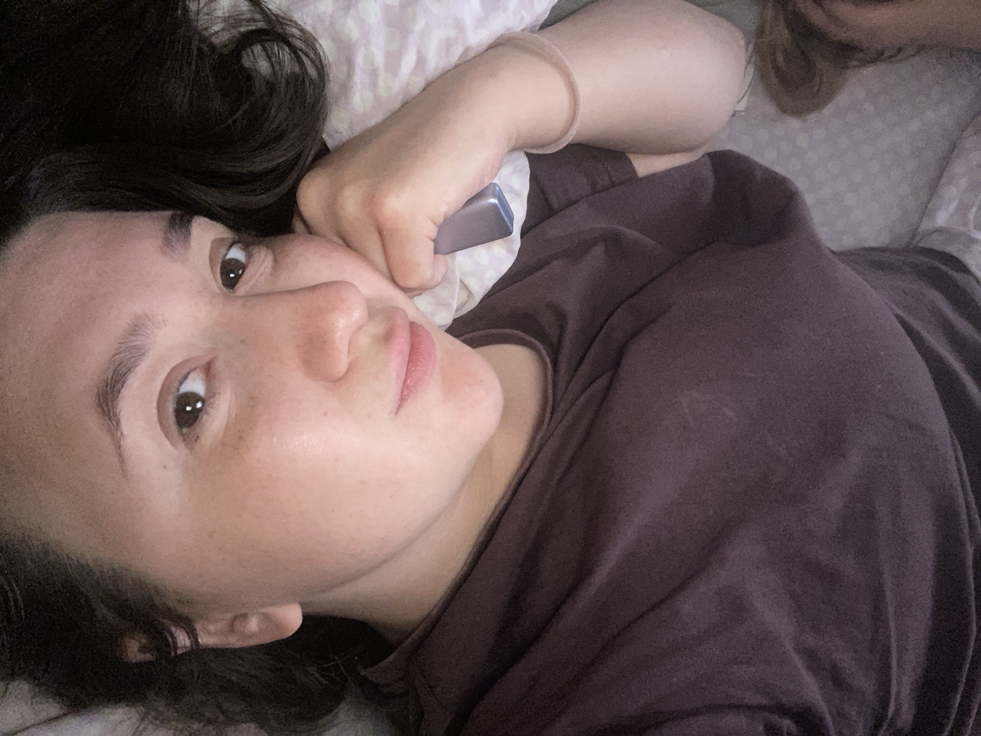 A woman with dark hair lies on a bed, holding a small, rectangular silver object to her face. She is wearing a brown t-shirt and a light-colored bracelet. The bed has a white pillow with a polka-dot pattern and a gray patterned sheet. A portion of another person's hair is visible in the upper right corner. The location is Chaikovsky, Russia.