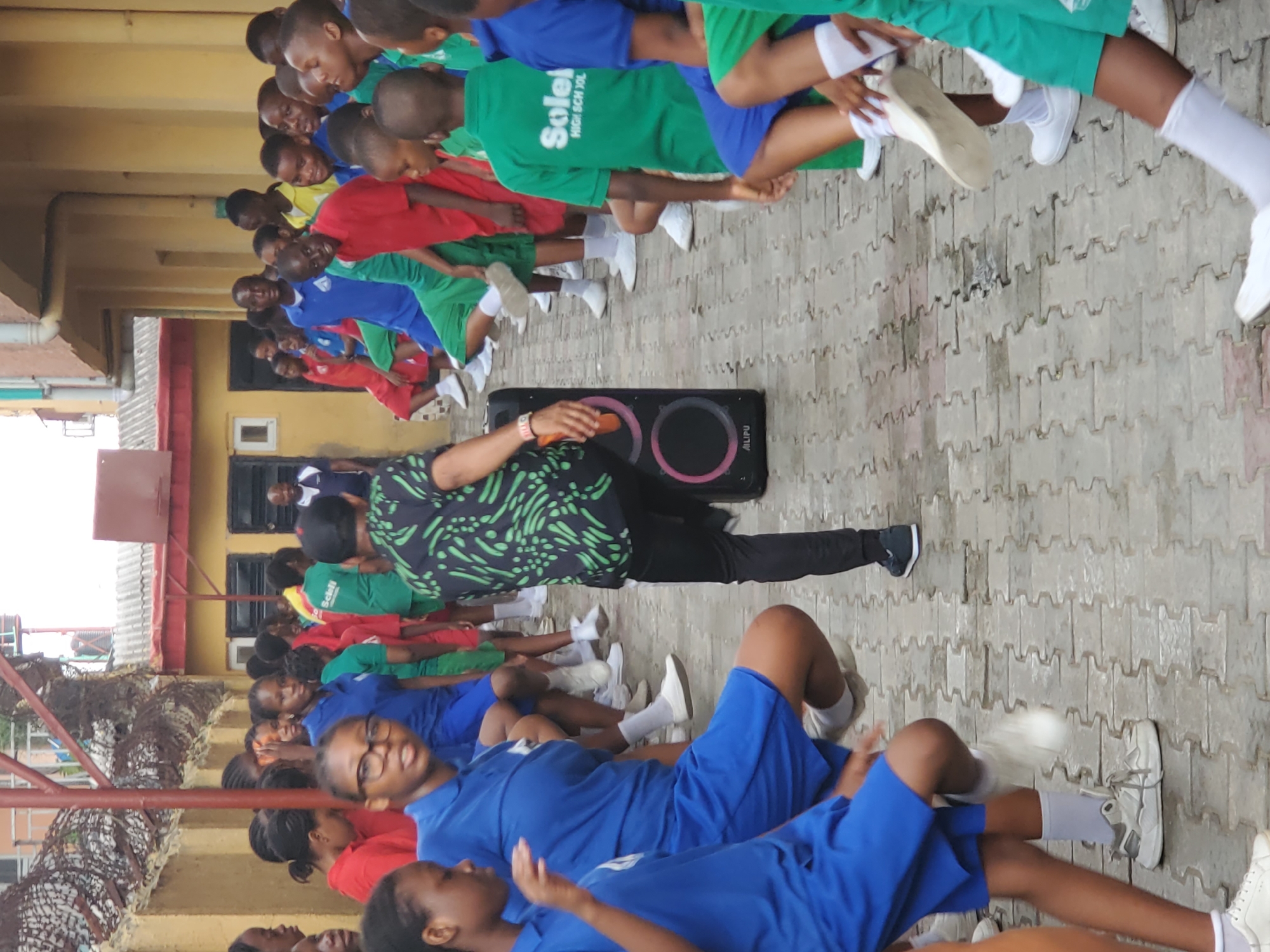 In Agani, Nigeria, a large group of school children are exercising outdoors on a paved surface near a building. The children are wearing school uniforms in colors of blue, red, and green. An adult male in a patterned shirt and black pants stands near a black speaker, appearing to lead the exercise. The children are lying on the ground doing a leg exercise while the adult faces them and is interacting with the speaker. A fence is visible in the foreground, and a building with a small window is in the background.
