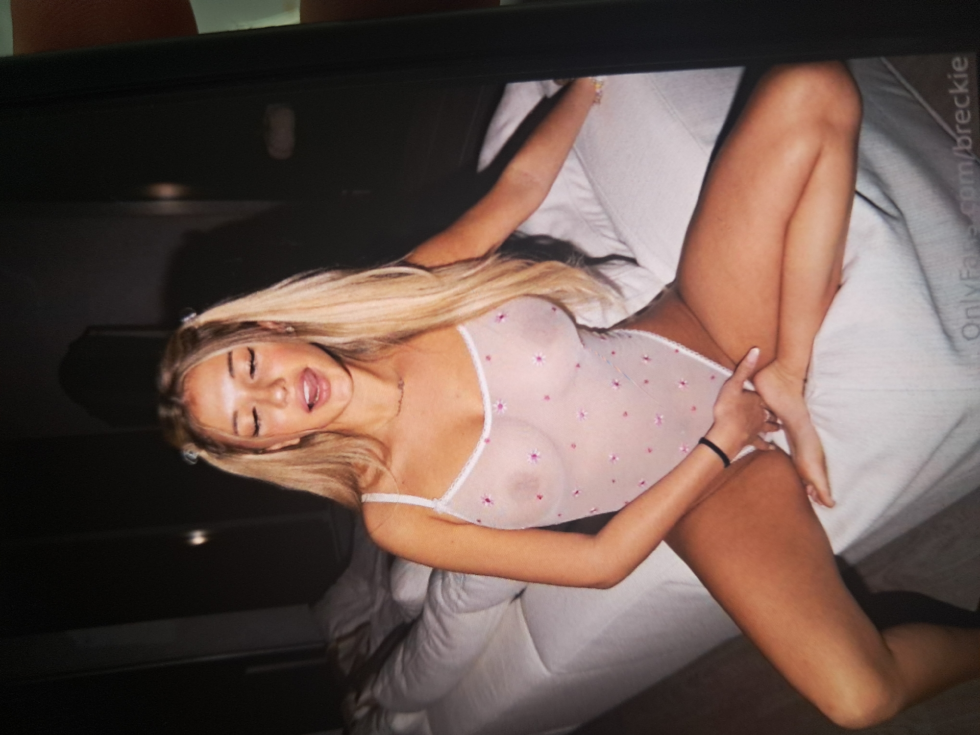 A young blonde female is depicted in an indoor, dimly lit setting. She is seated on a white upholstered surface, resembling a bed or large couch, with her legs slightly parted. Her long blonde hair is styled in two small pigtails on top of her head, with the remainder falling loosely. Her eyes are closed, and her mouth is open with her tongue slightly visible, conveying an expression of intense emotion.
She is wearing a sheer, light pink or white one-piece bodysuit with thin spaghetti straps, patterned with small pink or purple floral designs. Her nipples are visibly outlined through the translucent fabric. A delicate chain necklace is visible around her neck, and a black elastic hair tie is worn on her left wrist. Her left hand is placed between her thighs, resting near her inner thigh/groin area, while her right arm extends backward onto the white surface.
The background is dark and largely indistinct, suggesting a room with minimal lighting. No specific architectural features, landmarks, or details that would indicate a particular geographical location are visible within the image. A watermark displaying "OnlyFans.com/breckie" is present in the lower right corner of the image. The original image appears to be a photograph of a screen, with part of a dark frame visible on the right edge.