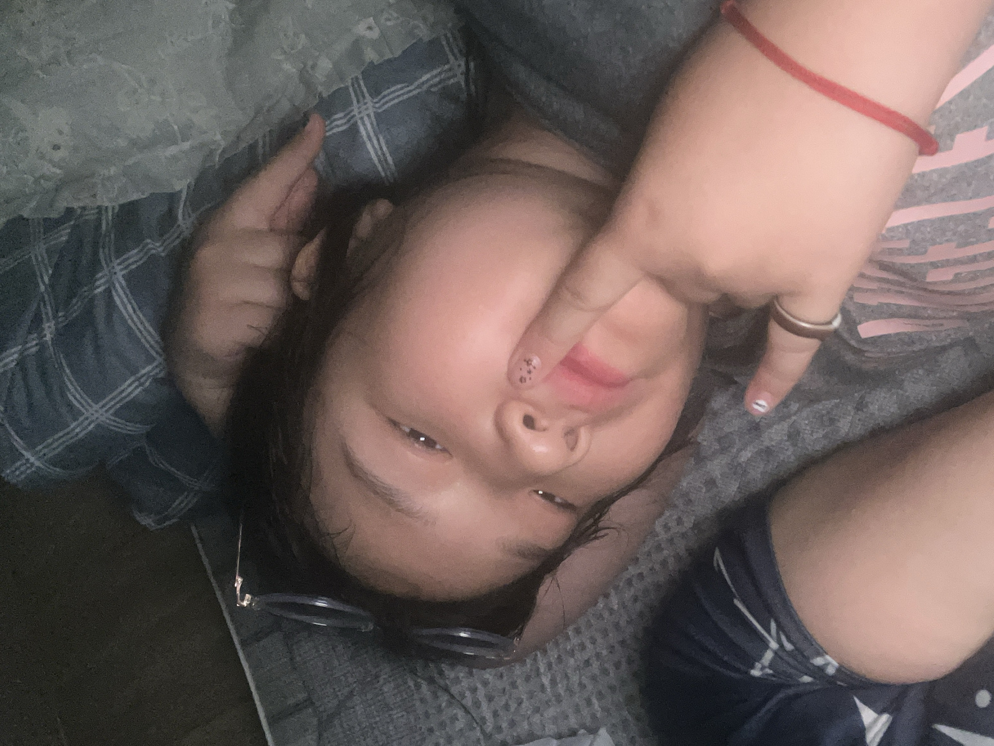 A person is lying on a bed with a hand covering part of their face. The bed has a plaid pattern. The person is wearing a gray shirt with pink stripes and a red bracelet on their wrist. They also have a ring on their finger and painted nails. A pair of glasses is on the bed near the person's head. The environment appears to be a bedroom. The city and specific location are unknown.
