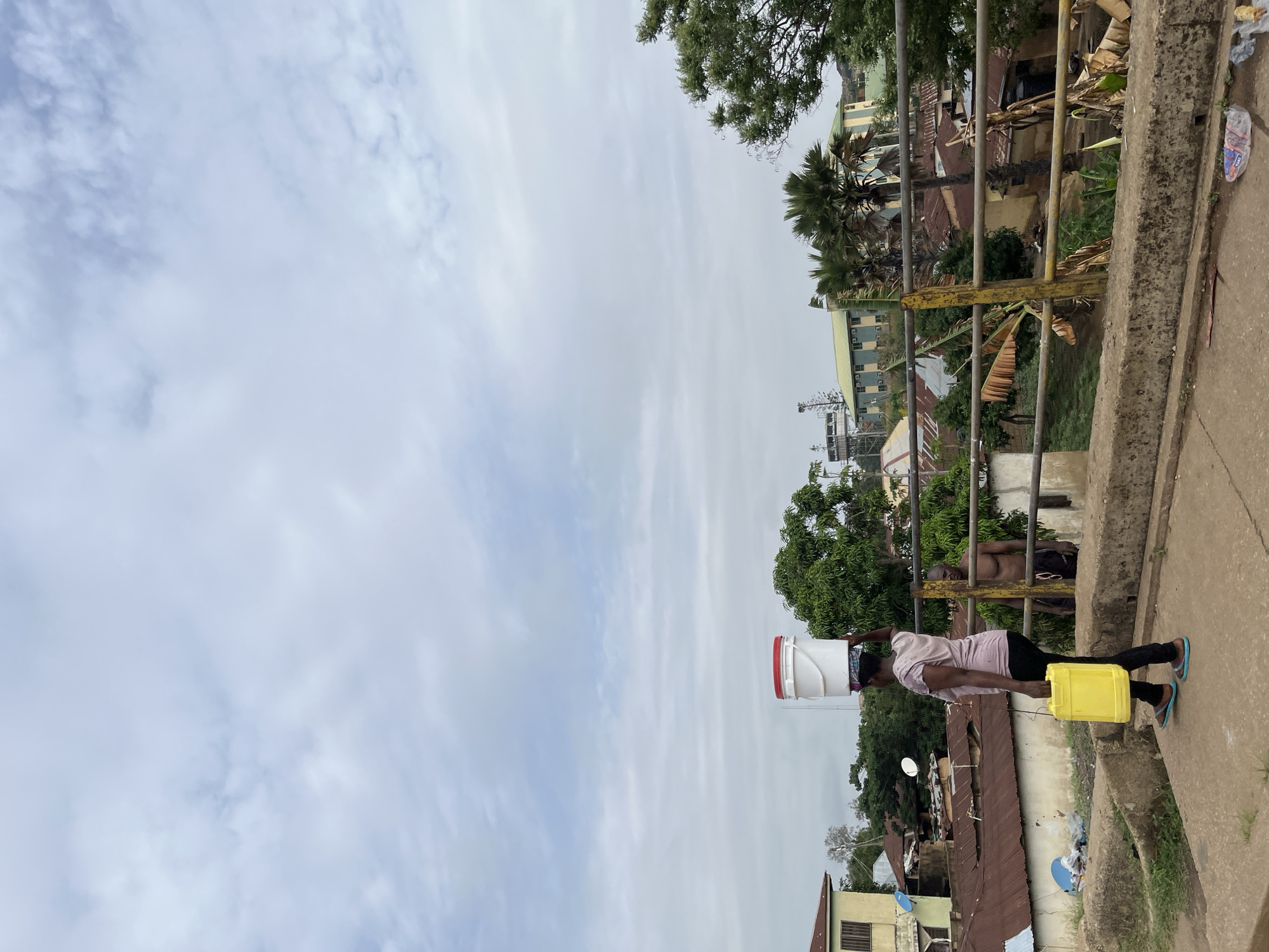 A woman stands on a concrete bridge in Jos, Nigeria, carrying a stack of white buckets on her head and a yellow container in her hand. She wears a light-colored shirt, dark pants, and blue sandals. Behind her, a shirtless man with sunglasses stands near the bridge railing. Houses with rusted metal roofs and lush green trees populate the background. The sky is overcast. A dilapidated yellow railing flanks the edge of the bridge.