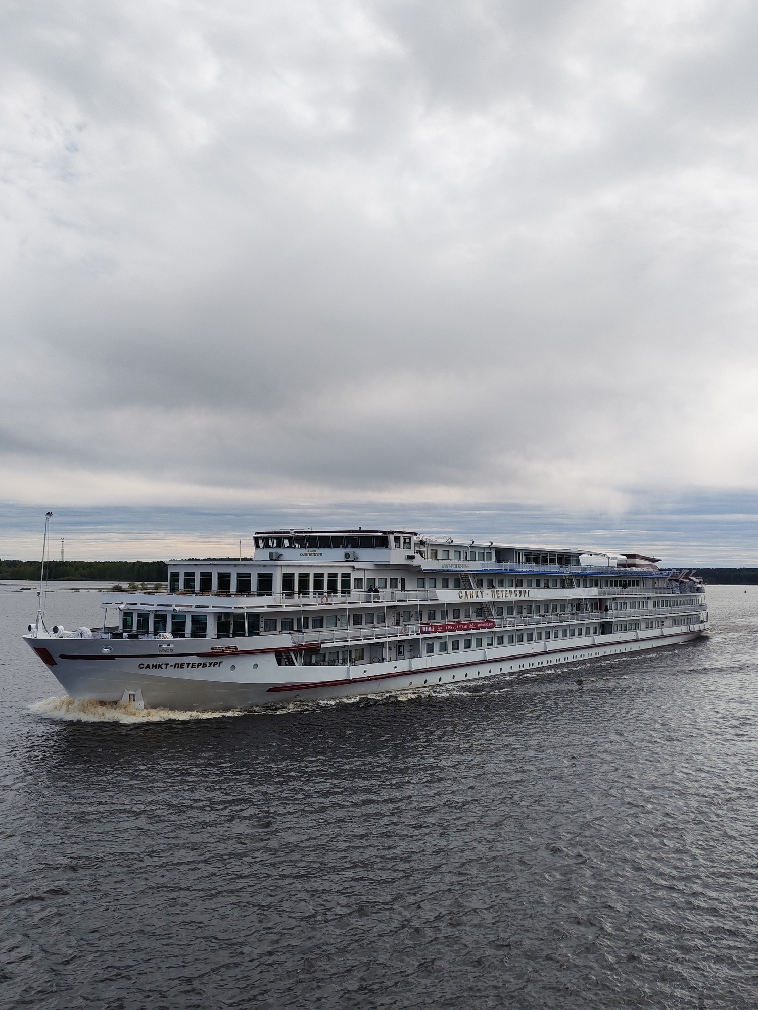 The image captures a large, multi-decked river cruise ship named "Sankt-Peterburg" moving on a body of water, possibly a river or lake. The ship is white with red trim, and the name is written in Cyrillic on its bow and side. The sky is overcast with grey clouds. In the background, a strip of land is visible, suggesting a shoreline. The water is dark and choppy, indicating some wind or current. The setting could be the city of Myshkin, Russia, as the ship's name and the environment are consistent with Russian river cruises. No people are clearly visible in the image. The focus is on the ship and its movement through the water.