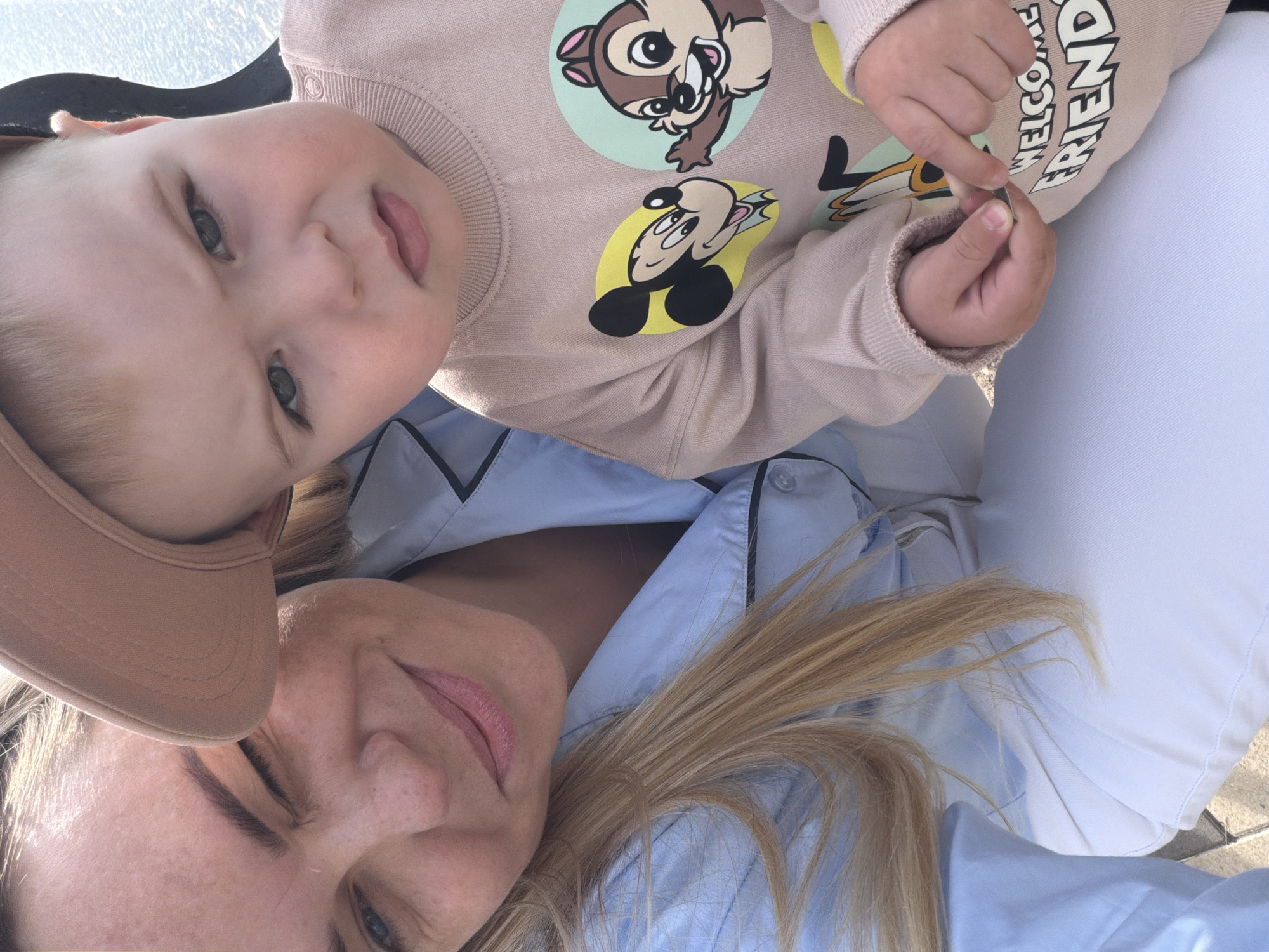 This close-up selfie shows a woman and a young child, likely a mother and her baby or toddler, bathed in bright sunlight indicating daytime outdoors. The image is taken from a slightly overhead perspective.
The woman is positioned in the lower part of the frame, looking up at the camera with a gentle smile. She has blonde hair, visible freckles on her face, and is wearing a light blue collared shirt with black trim. A beige cap is partially visible on her head.
Above the woman, the child is looking towards the camera with a calm expression and slightly parted lips. The child has light hair and is wearing a light beige long-sleeved sweatshirt. The sweatshirt features several cartoon characters, including Chip and Mickey Mouse, depicted in colorful circular motifs. Visible text on the child's right sleeve reads "WELCOME FRIEND." The child's hands are clasped together, holding a small, indistinct object.
The background is largely out of focus, showing bright light and blurred shapes, suggesting an outdoor setting. A portion of white fabric, possibly pants, is visible next to the child. The overall mood is warm and tender, capturing an intimate moment between the two.