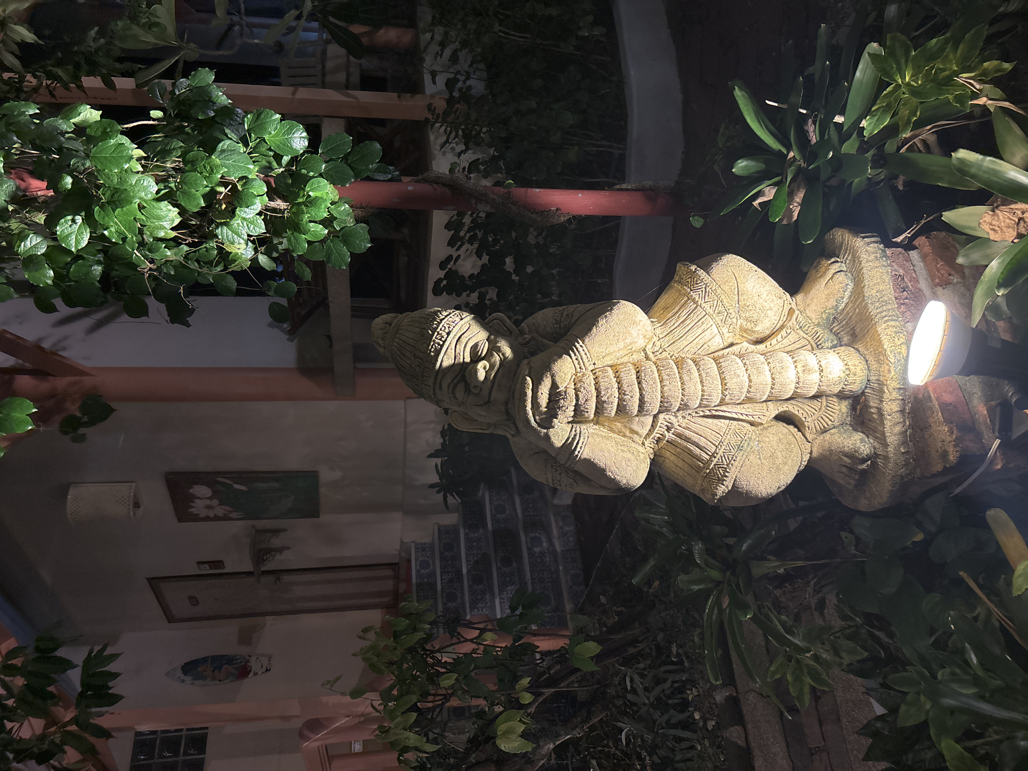 An outdoor, low-angle, nighttime shot captures a stone statue of a mythical guardian figure in the foreground. The statue, weathered and textured, is illuminated by a low-mounted spotlight. It is depicted in a seated, contemplative pose, with its head resting on its clasped arms, which are wrapped around its chest. The figure wears a conical hat and ornate, layered clothing. The statue sits on a brick platform at the base of a tree with lush green foliage. Behind the statue, a set of outdoor stairs with decorative tiles ascends to an entrance. The entrance features a closed door, a wall-mounted light fixture, and two pieces of artwork on the wall: a small, round depiction of figures and a larger, rectangular painting of flowers. The environment appears to be a well-maintained courtyard or garden area, possibly part of a hotel or residence, located in Chalong, Thailand. The overall scene is dark, with the lighting highlighting the statue and some of the surrounding vegetation, creating a serene and somewhat mysterious atmosphere.