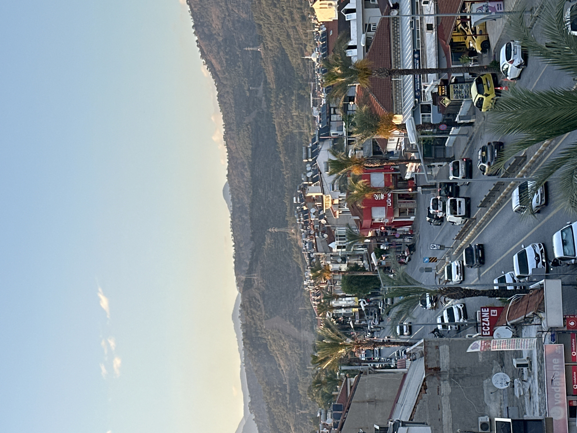 The image shows a view from above, likely a balcony or high building, overlooking a street in Fethiye, Turkey. The street is lined with buildings, some of which are commercial establishments indicated by signs in Turkish such as "ECZANE" (pharmacy) and advertisements for "Vodafone." Palm trees are planted along the street, adding to the coastal town atmosphere. Vehicles, including cars, taxis (some yellow), and vans, are visible on the road, suggesting traffic. In the background, a mountain range rises, providing a scenic backdrop to the urban landscape. The sky is clear, indicating daytime. The general impression is of a vibrant, busy street in a tourist area within Fethiye.