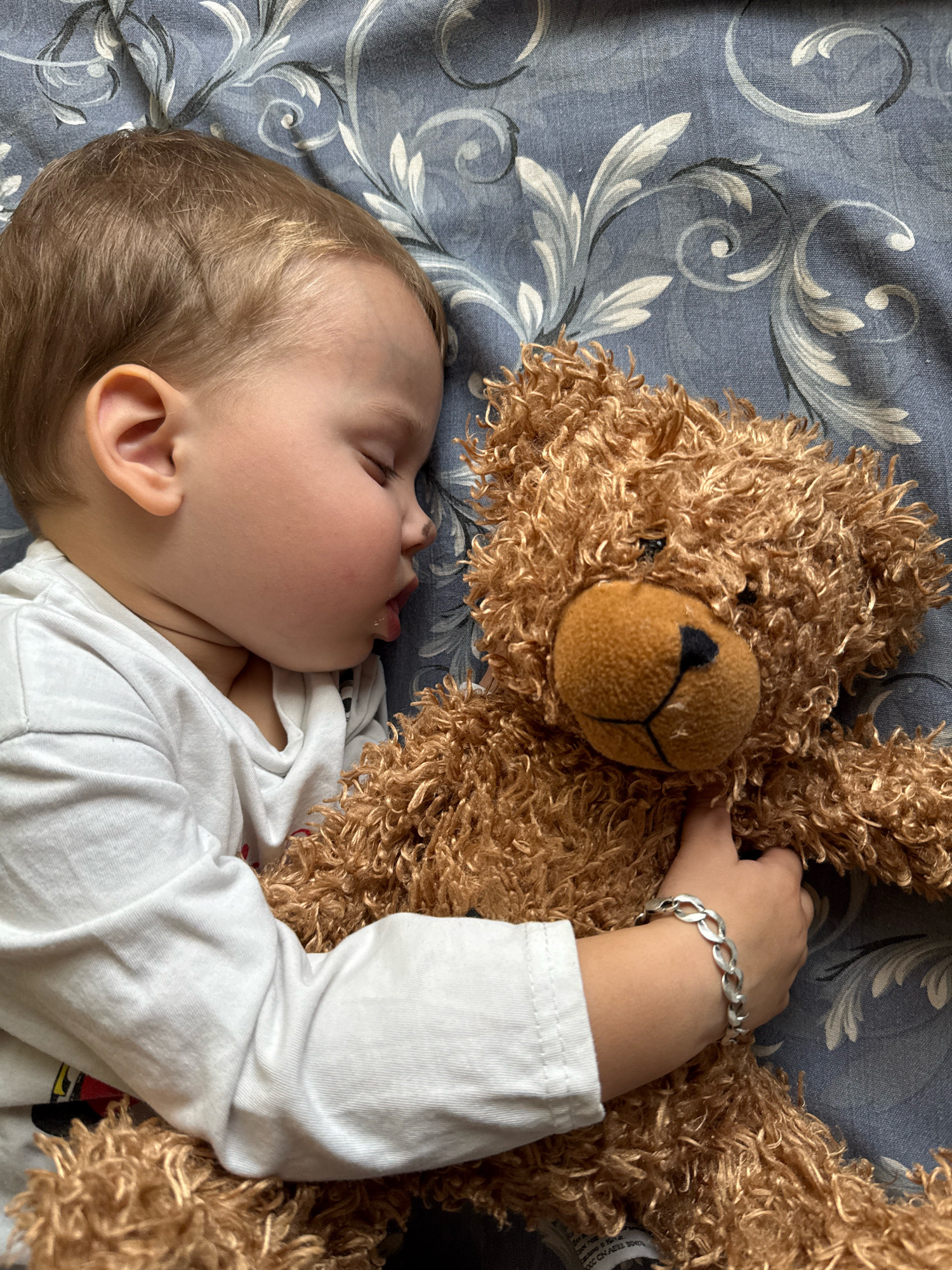A young child with blond hair and closed eyes is sleeping soundly, cuddled with a brown teddy bear. The child is wearing a white long-sleeved shirt. The teddy bear is a light brown color with curly fur and is being held by the child's arms. The child's left wrist has a silver bracelet on it. They are lying on a bed with blue patterned sheets. The scene appears to be indoors, possibly in a bedroom in the city of Rusanovo, Russia.