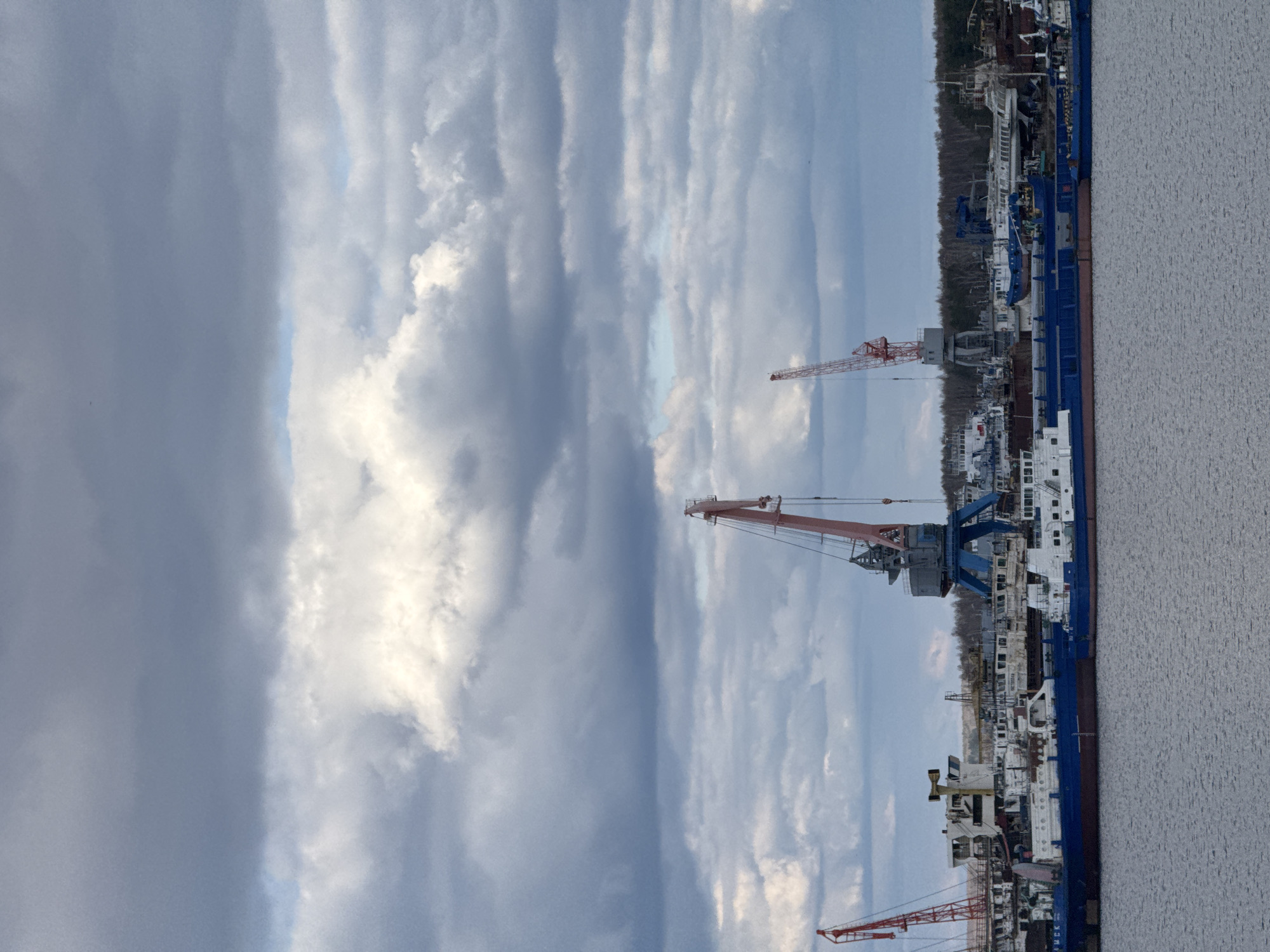 The image captures an active river port or shipyard in Chaikovsky, Russia, under a dramatic sky. The bottom third of the frame is dominated by a calm expanse of rippled, greyish water. Along the opposite bank, several large industrial ships or barges are docked. These vessels are predominantly blue and white, with some reddish-brown hulls visible, suggesting they are cargo ships or undergoing maintenance.
Numerous tall, red-and-white industrial cranes are prominently positioned along the dock, indicating ongoing loading, unloading, or construction activities. Various industrial buildings, structures, and scaffolding are visible lining the bank behind the vessels. A dense line of bare trees forms a natural backdrop to the industrial complex, hinting at late autumn, winter, or early spring.
The upper half of the image is filled with a vast sky, characterized by long, streaky clouds in varying shades of grey and white. Patches of lighter blue sky are visible between the cloud formations, suggesting a dynamic weather pattern. The lighting is diffused, consistent with an overcast day or early morning/late afternoon. On the blue hull of one of the ships, faint Cyrillic text, possibly "ЧЕЛНЕК" (CHELNEK), can be seen. The scene depicts typical industrial maritime operations at a bustling waterway hub.