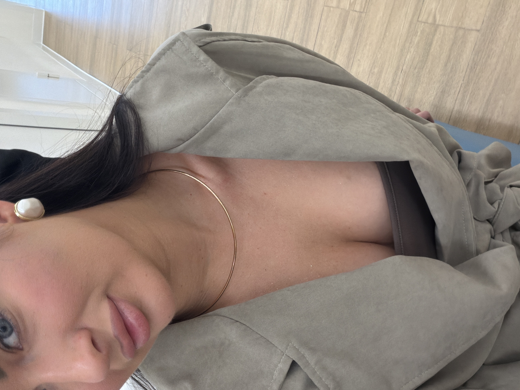 A close-up photograph depicts a woman with fair skin, dark hair, and blue eyes. She is wearing a gold necklace and a beige jacket, partially open at the chest. A single earring is visible. The background consists of wooden flooring and a white object resembling furniture. The location and city are not deducible from the image.